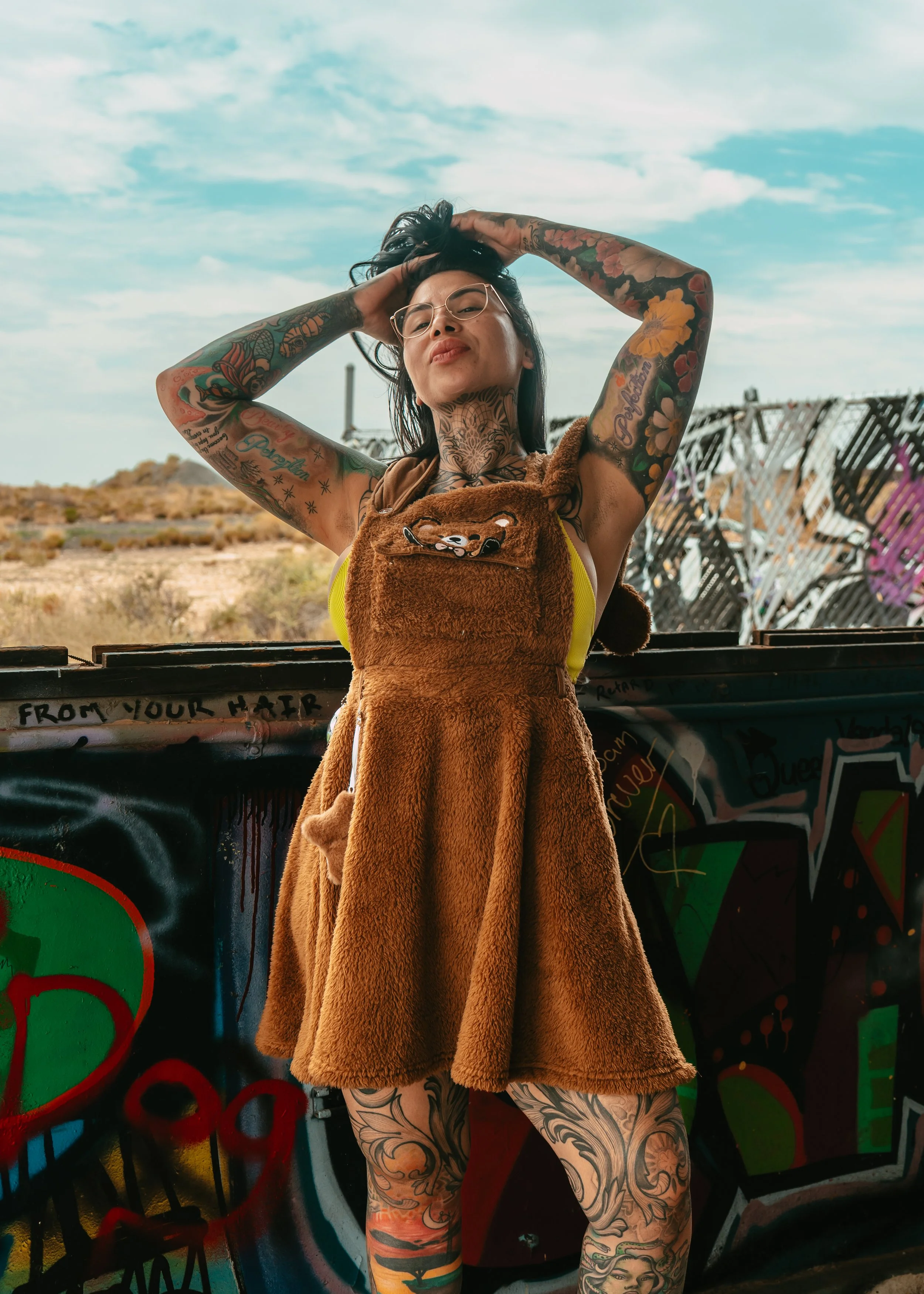 A tattooed woman with glasses stands outdoors with arms raised, wearing a bear-themed fuzzy dress and glasses. Behind her is a graffitied wall and a landscape with a partly cloudy sky.