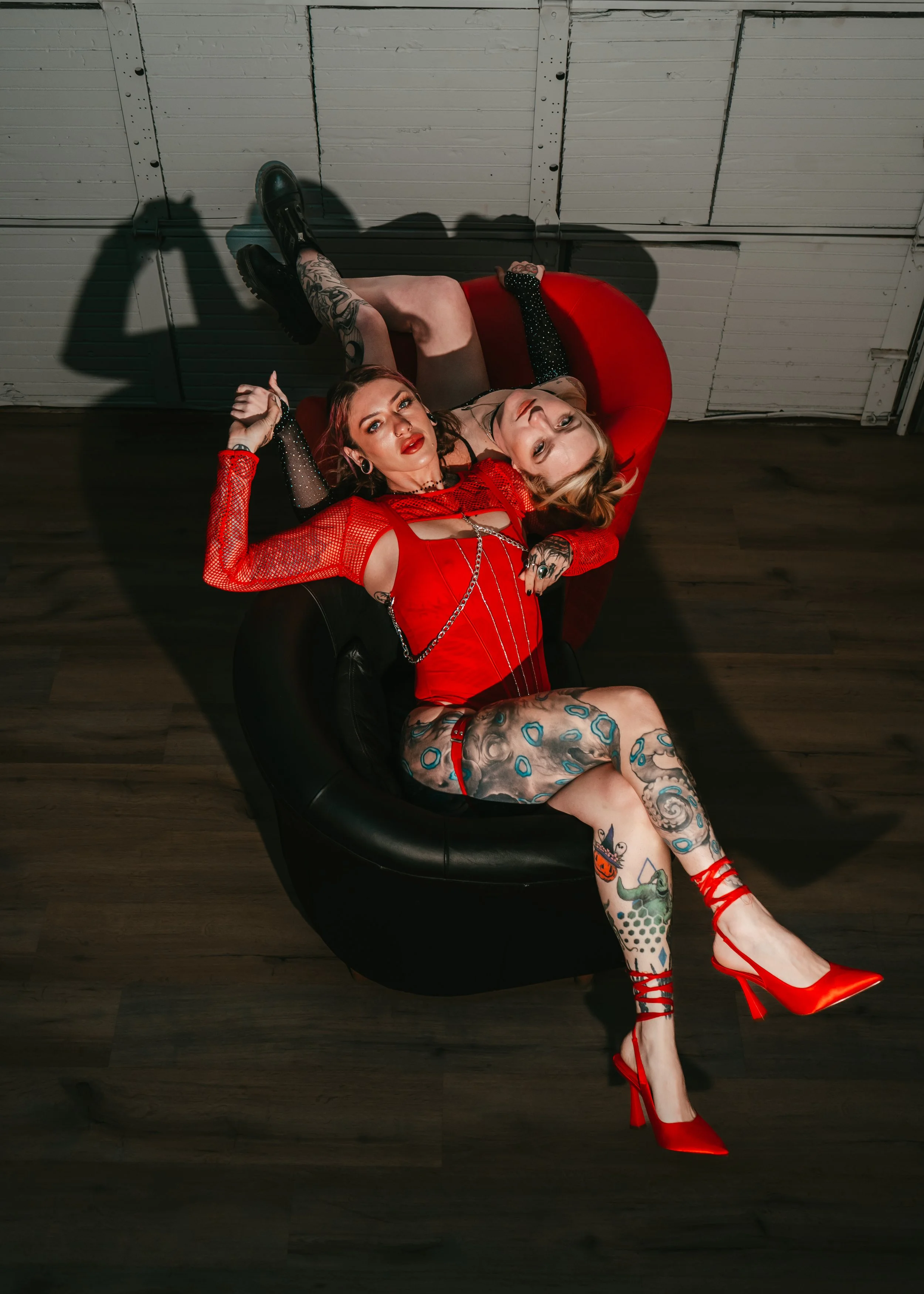 Two women with tattoos and dark makeup lying on a black and red couch, above a wooden floor, in a room with a white paneled ceiling.