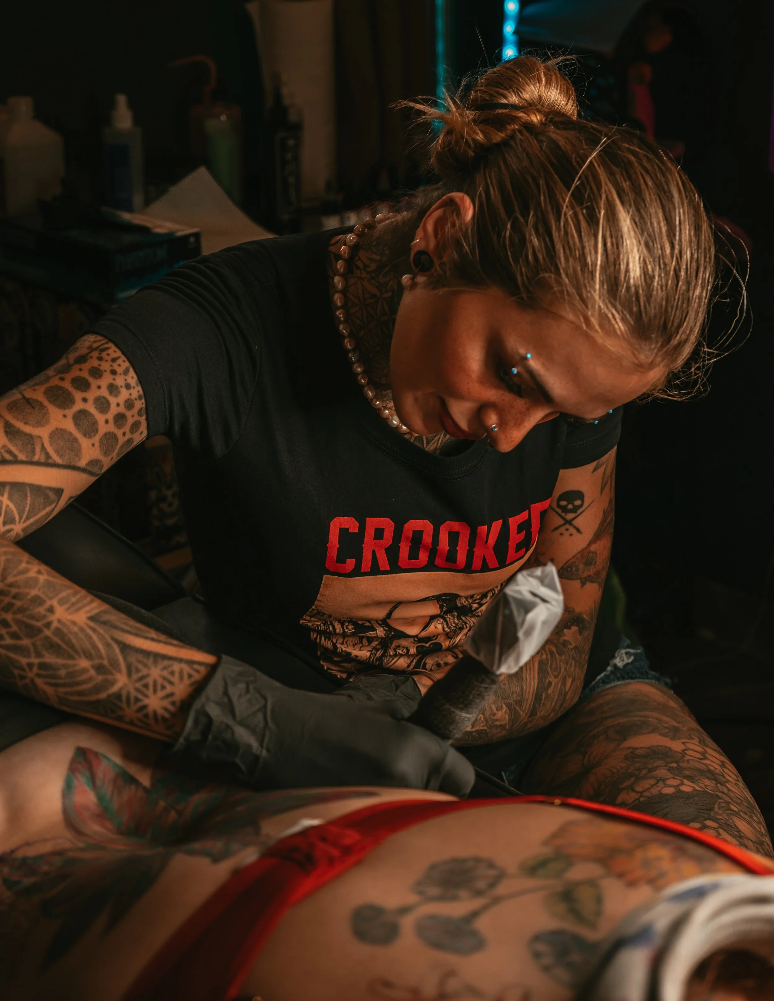 A woman with tattoos and piercings, wearing a black T-shirt with the word 'CROOKED,' tattooing a person's thigh in a tattoo studio.