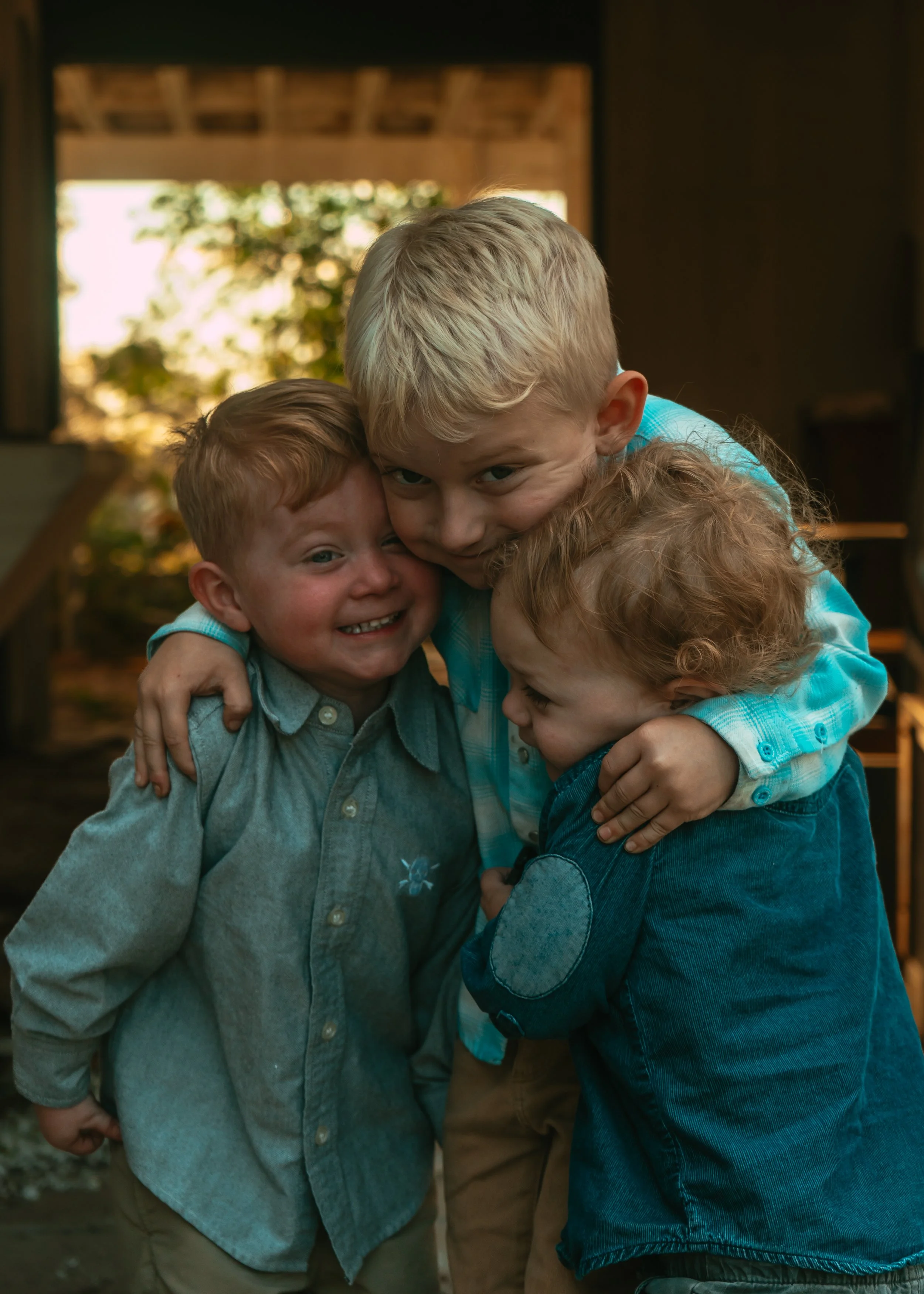 Four children hugging and smiling inside a wooden structure with sunlight and greenery in the background.