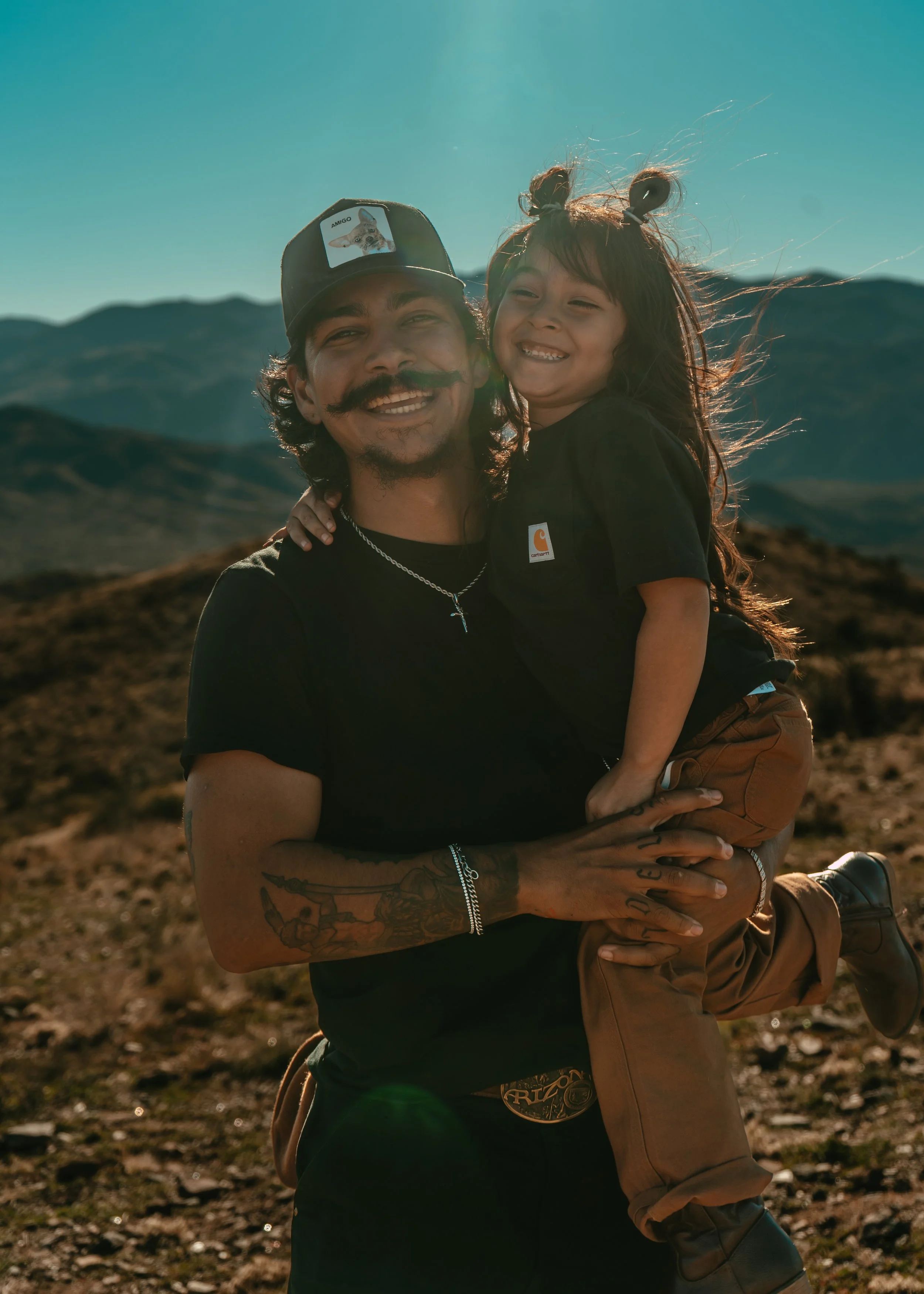 A man with long hair and a mustache, wearing a cap and black T-shirt, is smiling while holding a young girl in front of a mountainous landscape during daytime. The girl is smiling, has long hair styled in buns, and is wearing a black T-shirt and brown pants.