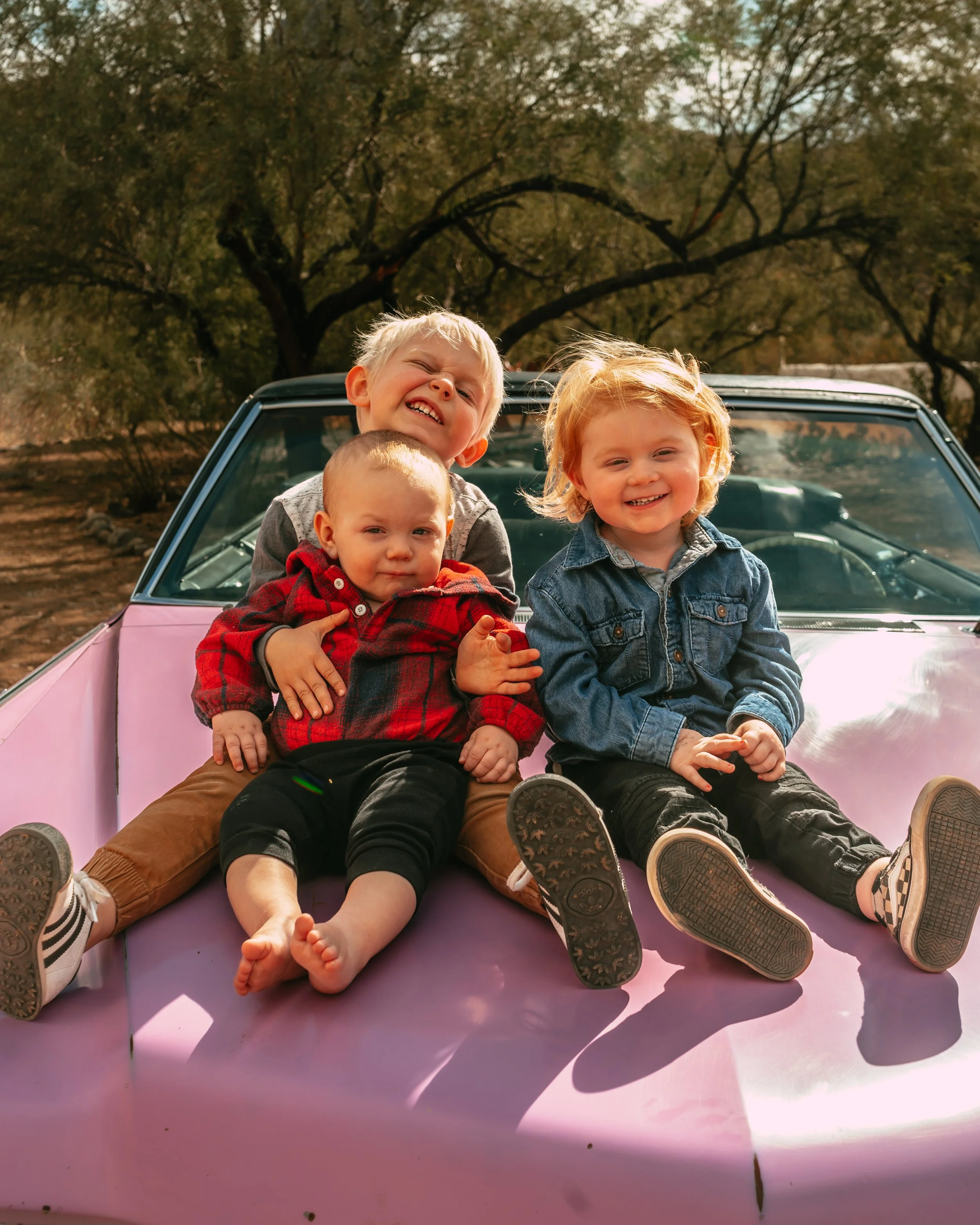 Three children sitting on the hood of a purple car outdoors, smiling and enjoying the moment.