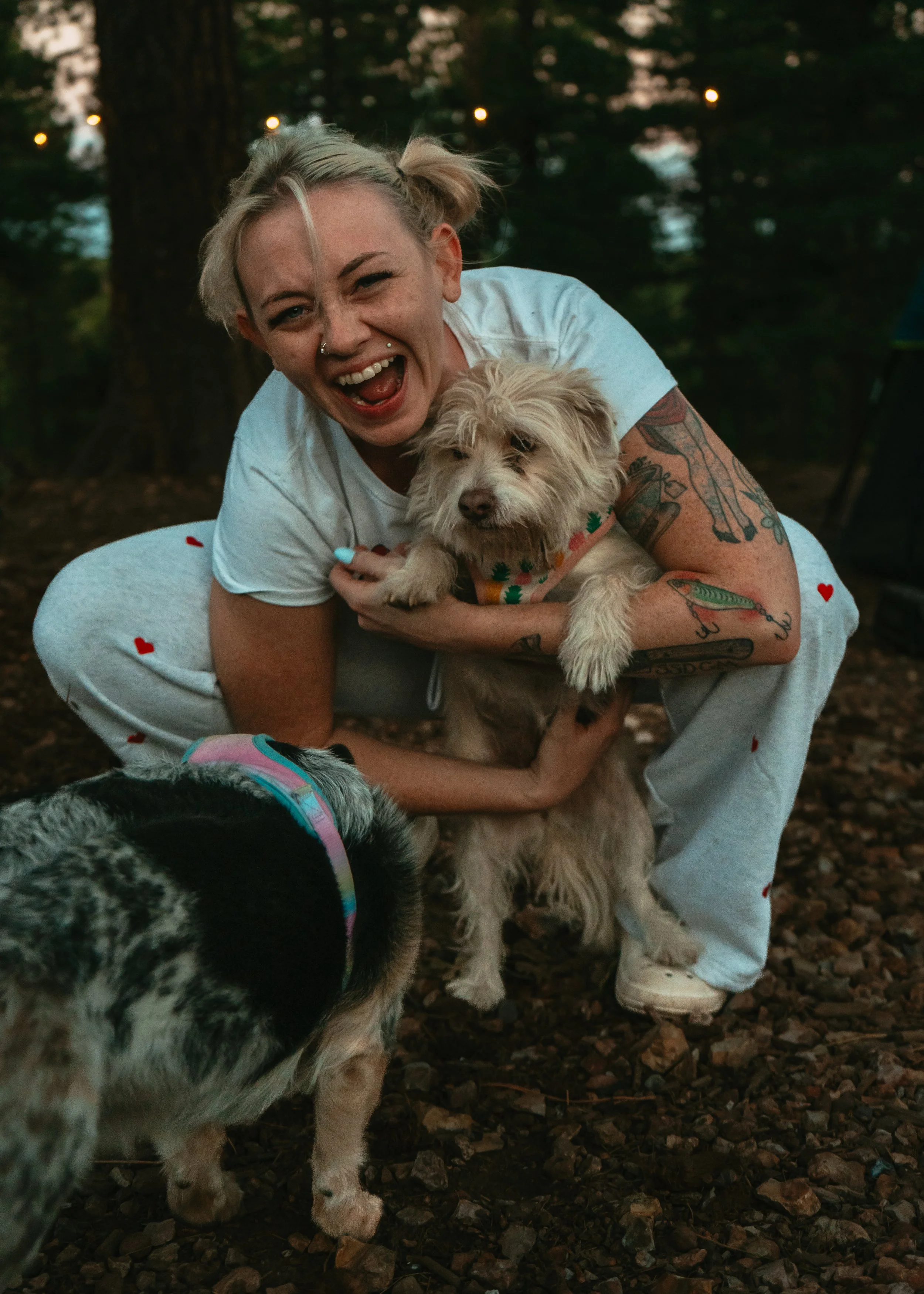 A woman with tattoos and multiple facial piercings smiling and holding a tan and white dog outdoors in a wooded area, with another dog partially visible in the foreground.