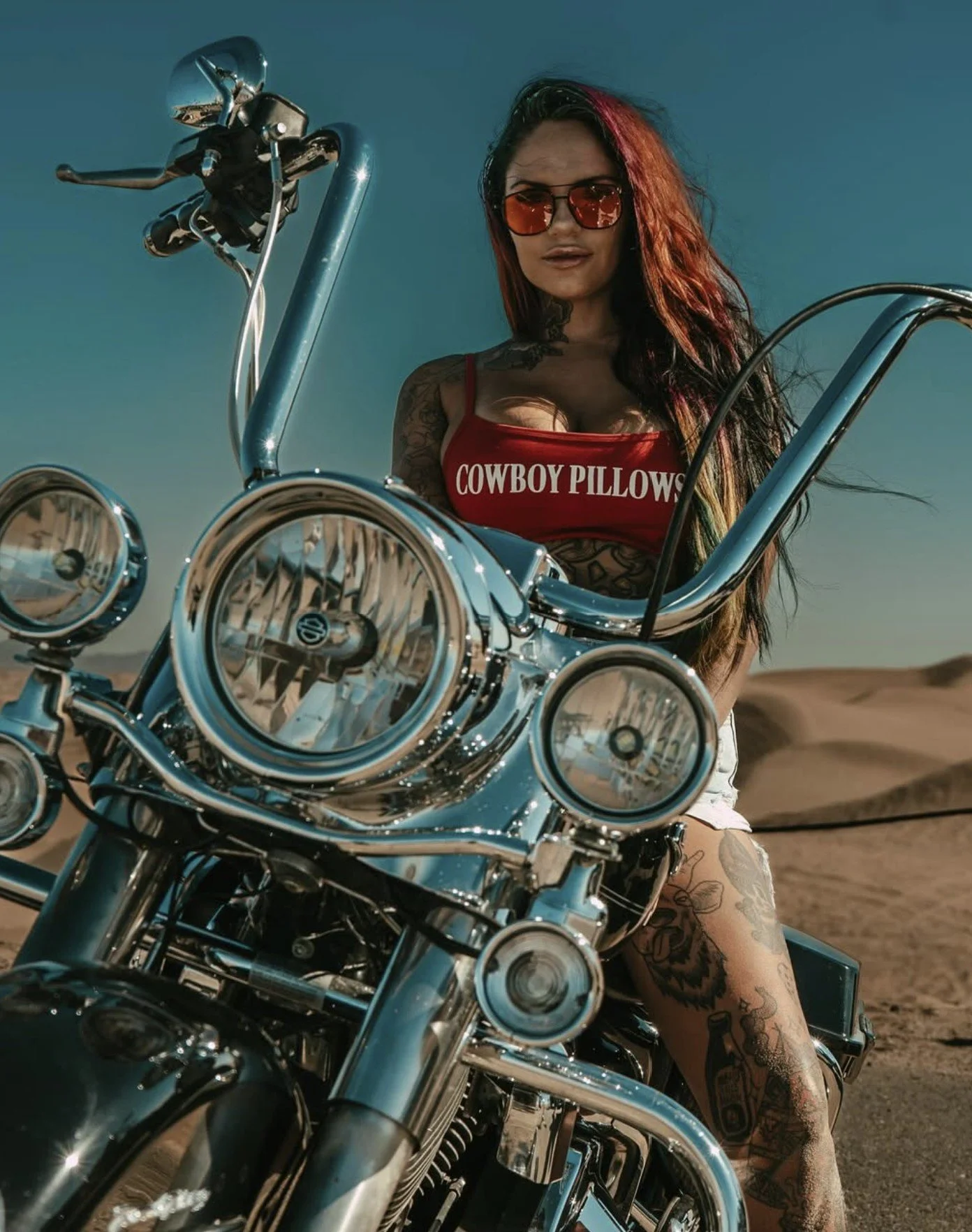 A woman with long hair and tattoos wearing red sunglasses and a red tank top that says 'Cowboy Pillows' sitting on a shiny chrome motorcycle in a desert landscape.
