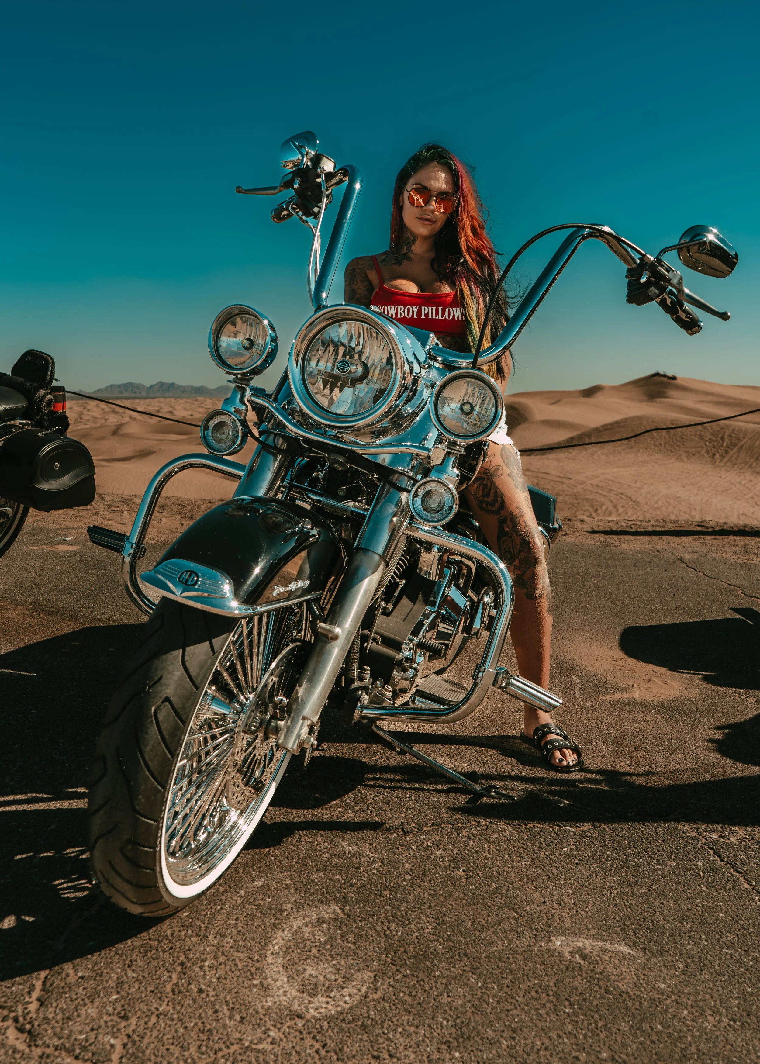 Woman with tattoos in sunglasses sits on a chrome motorcycle in desert landscape with sand dunes and clear blue sky.