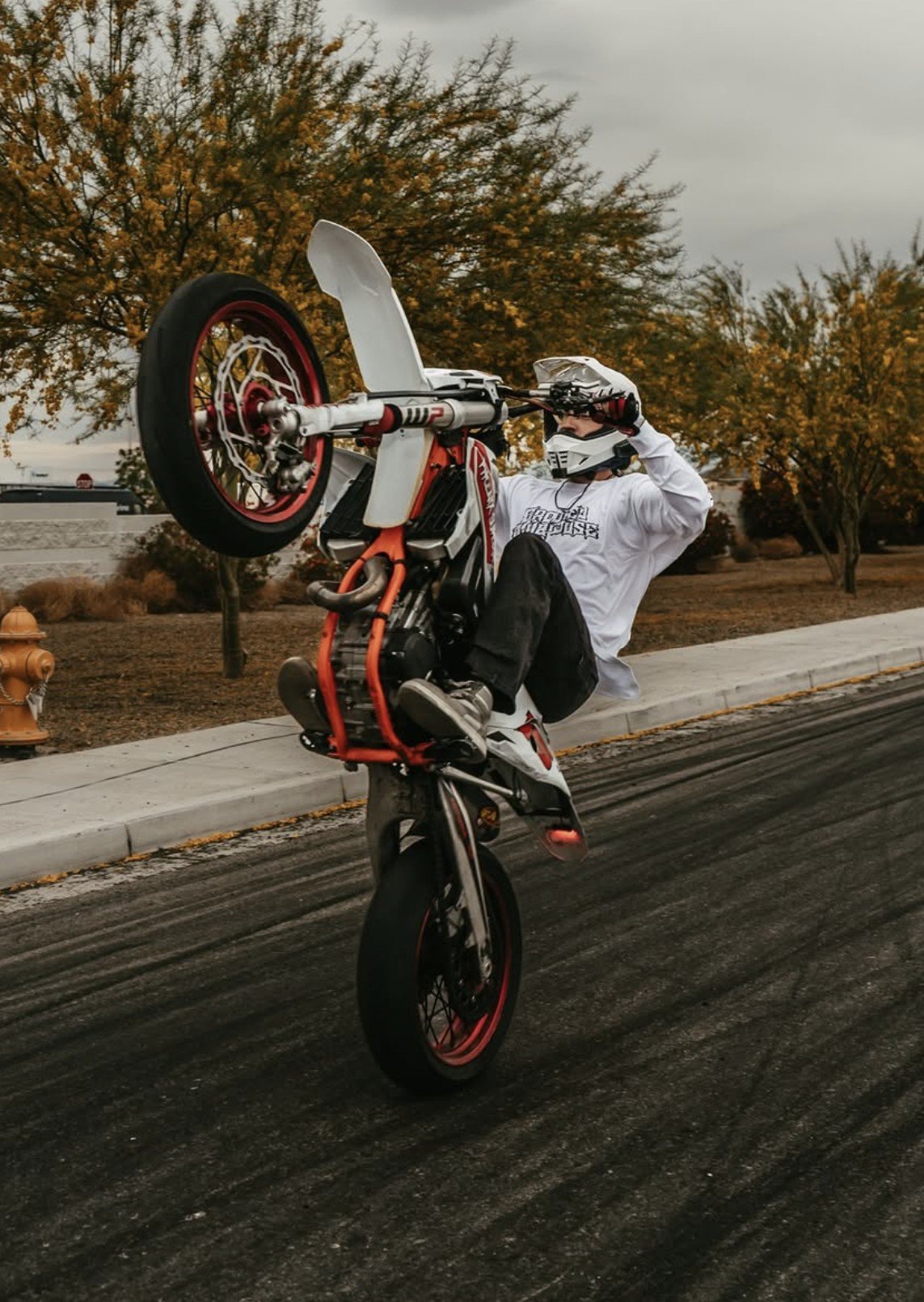 A person performing a wheelie on a dirt bike on a street with trees in the background.