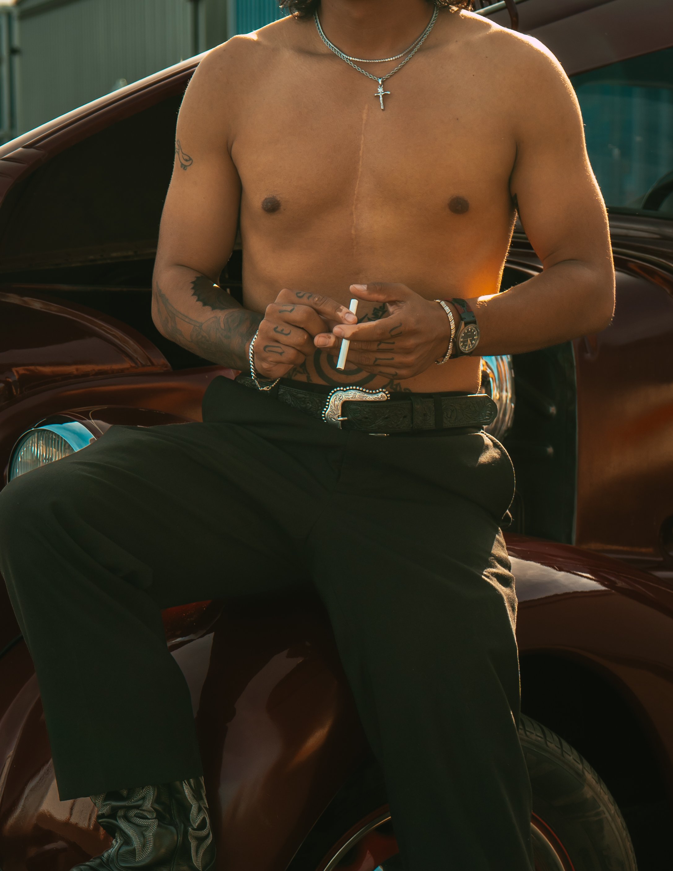 A shirtless man with tattoos on his arms and torso, wearing black pants, a belt, and cowboy boots, sitting on the hood of a vintage car, holding a cigarette, with a cross necklace, watch, and bracelets.