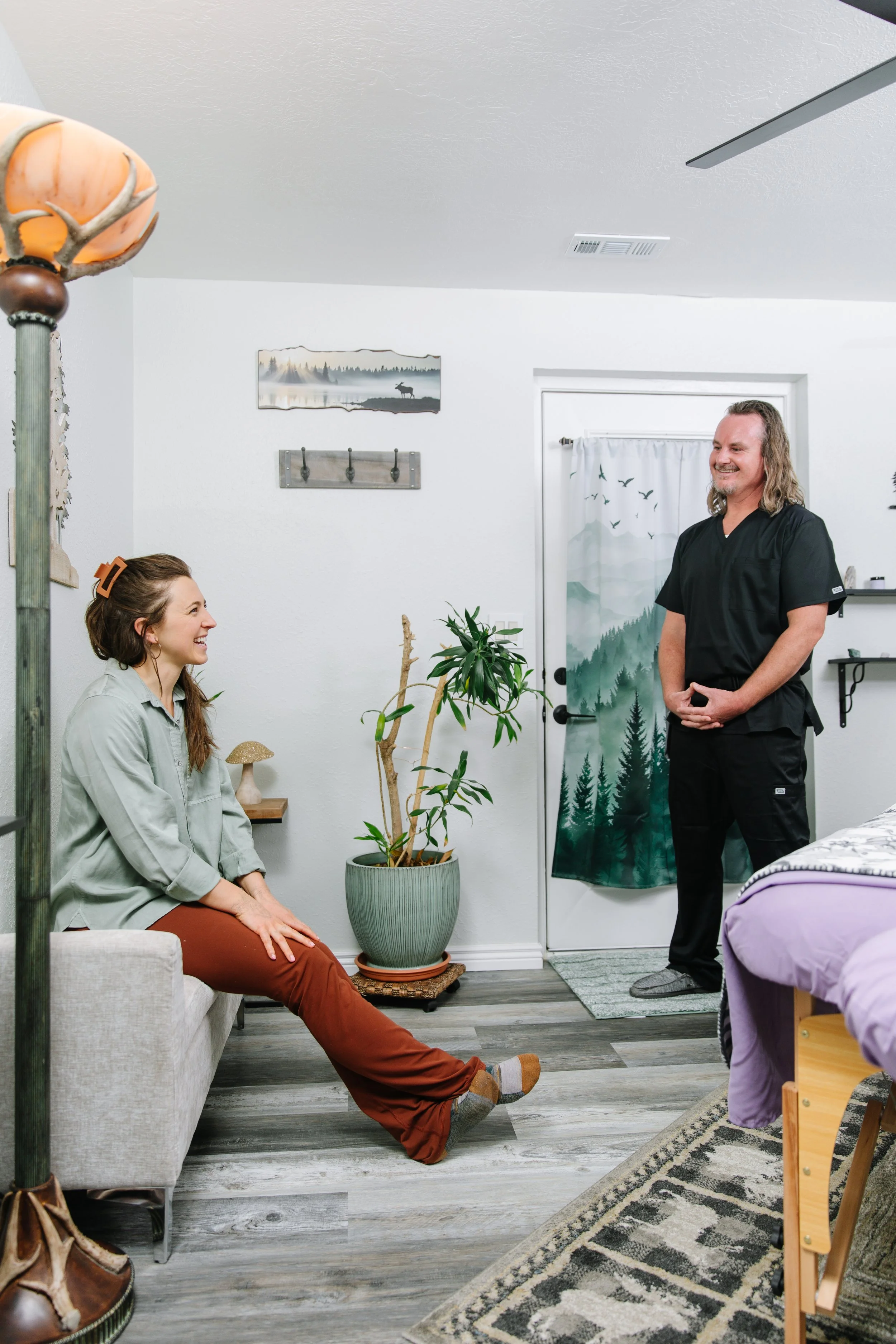 A woman sitting on a couch, smiling and talking to a man standing near a door, in a cozy room with houseplants, wall art, and a bed with purple bedding.