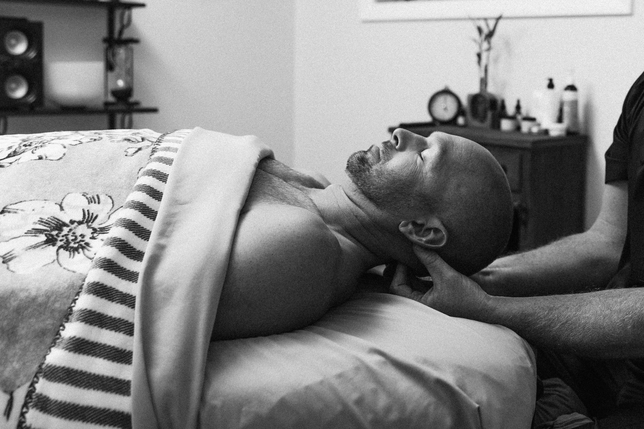 A man lying on his back on a bed with his eyes closed, receiving a neck massage from a person with their hands on his neck, in a bedroom setting.