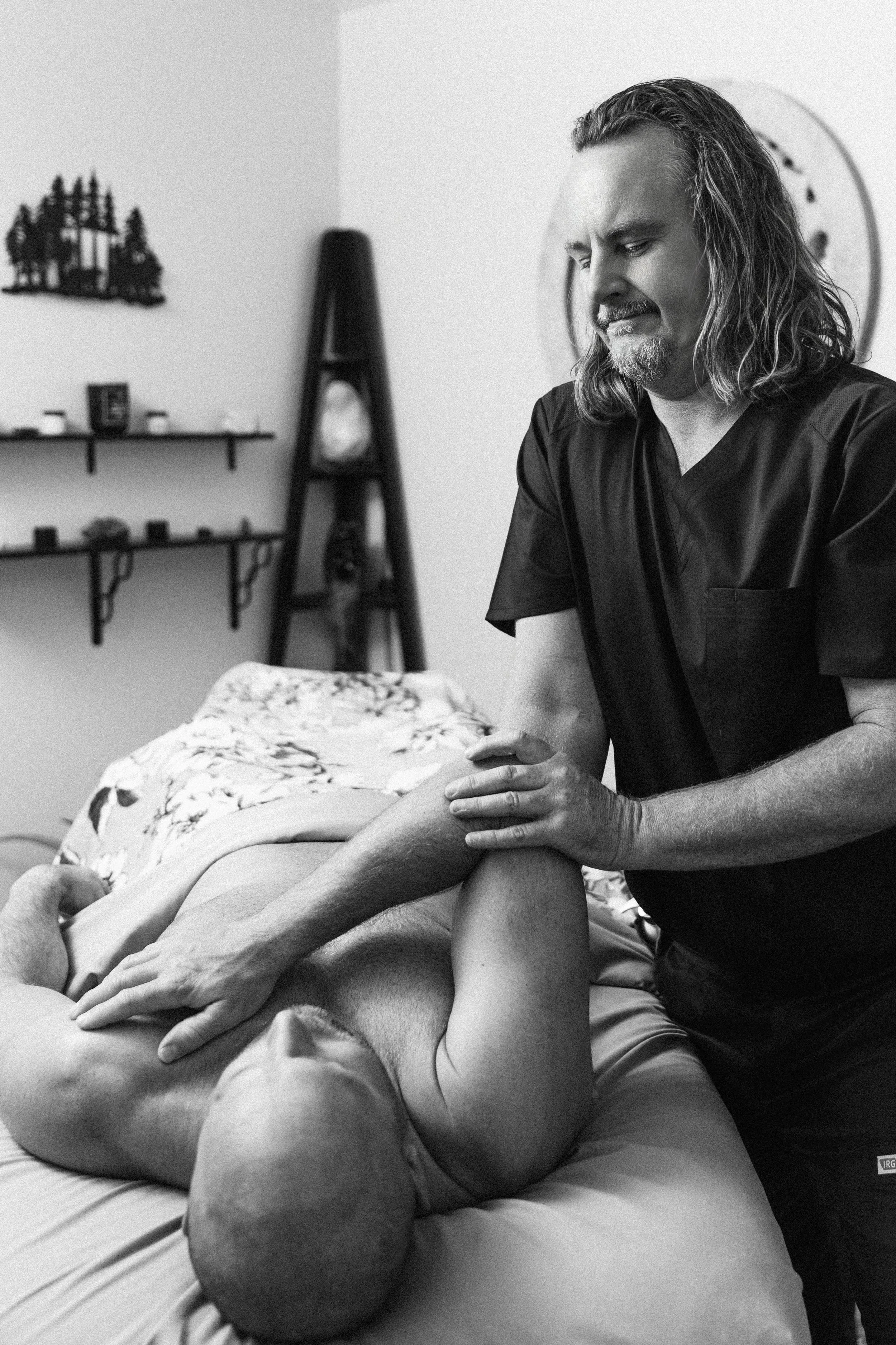A male massage therapist with long hair and a beard performs a massage on a client's arm in a massage room.