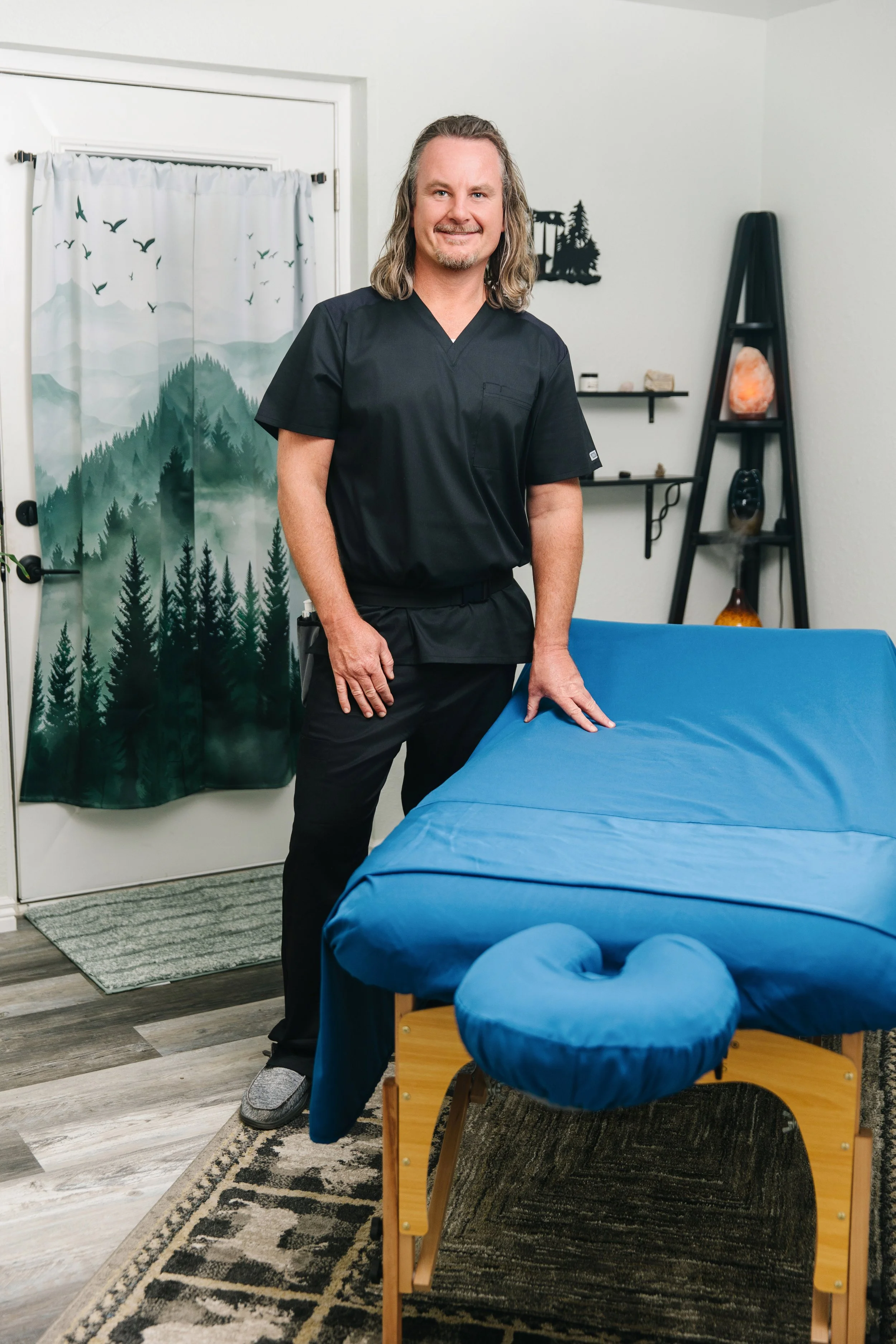 A man with shoulder-length hair and a beard, dressed in black scrubs, standing in a room with a massage table covered with a blue sheet and a heart-shaped cushion. The room has a door with a forest-themed curtain, wall shelves with rocks and decorative items, and a black ladder-style shelf with objects.
