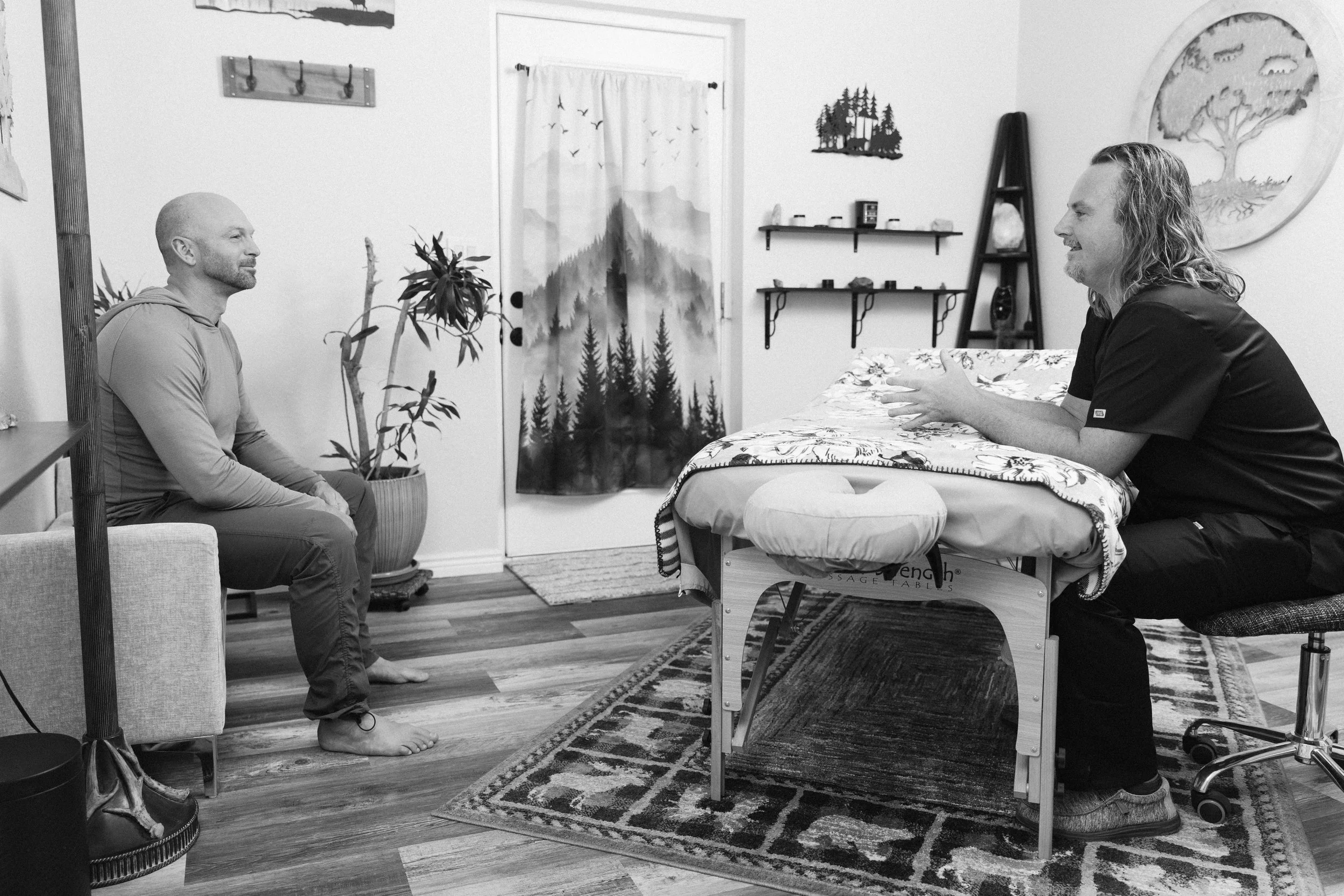A therapy session with two men. One man is sitting on a sofa, listening, while the other man is sitting on a stool, explaining something, in a room decorated with nature-themed artwork and shelves.