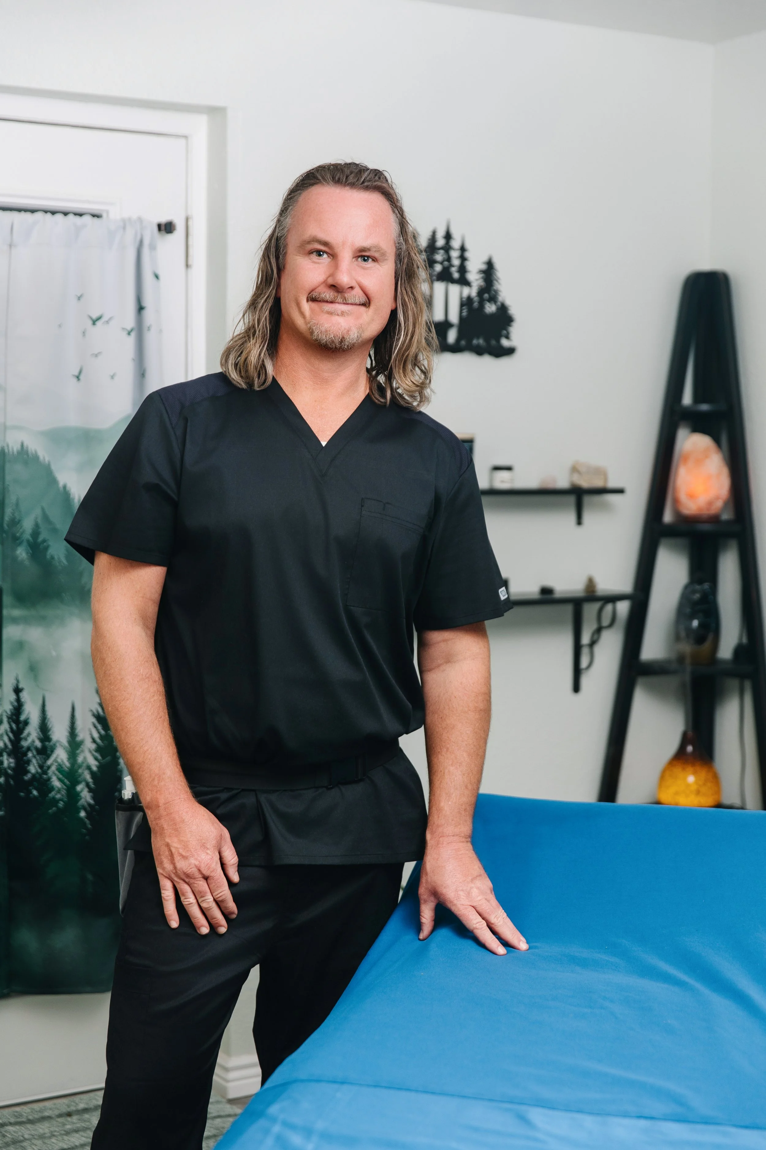 A man with long, wavy hair, wearing black medical scrubs, standing next to a blue table in a wellness or massage room with decor including a Himalayan salt lamp and nature-themed pictures.