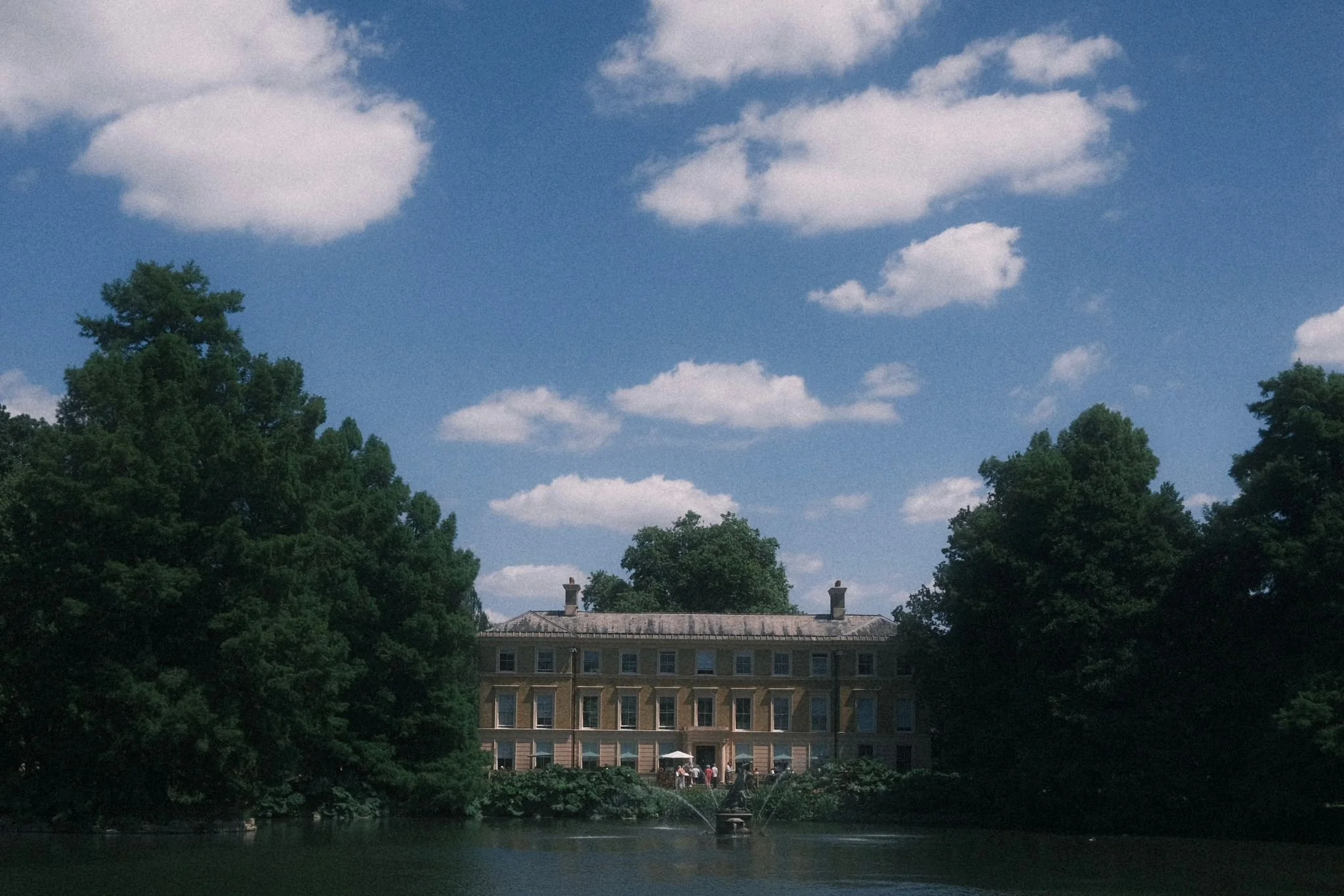 A large historic building with multiple windows seen through trees over a pond, with a fountain and people gathered outside. The sky is blue with scattered white clouds.