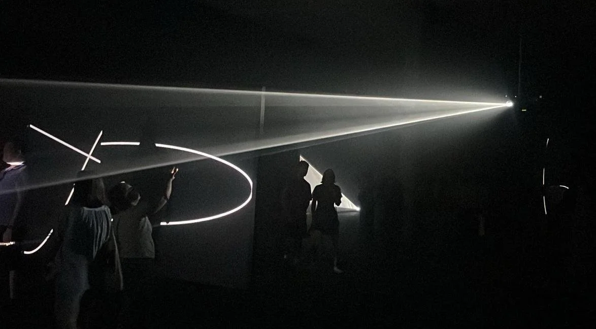 People walking through a dark indoor space with illuminated light trails and laser beams creating geometric patterns.