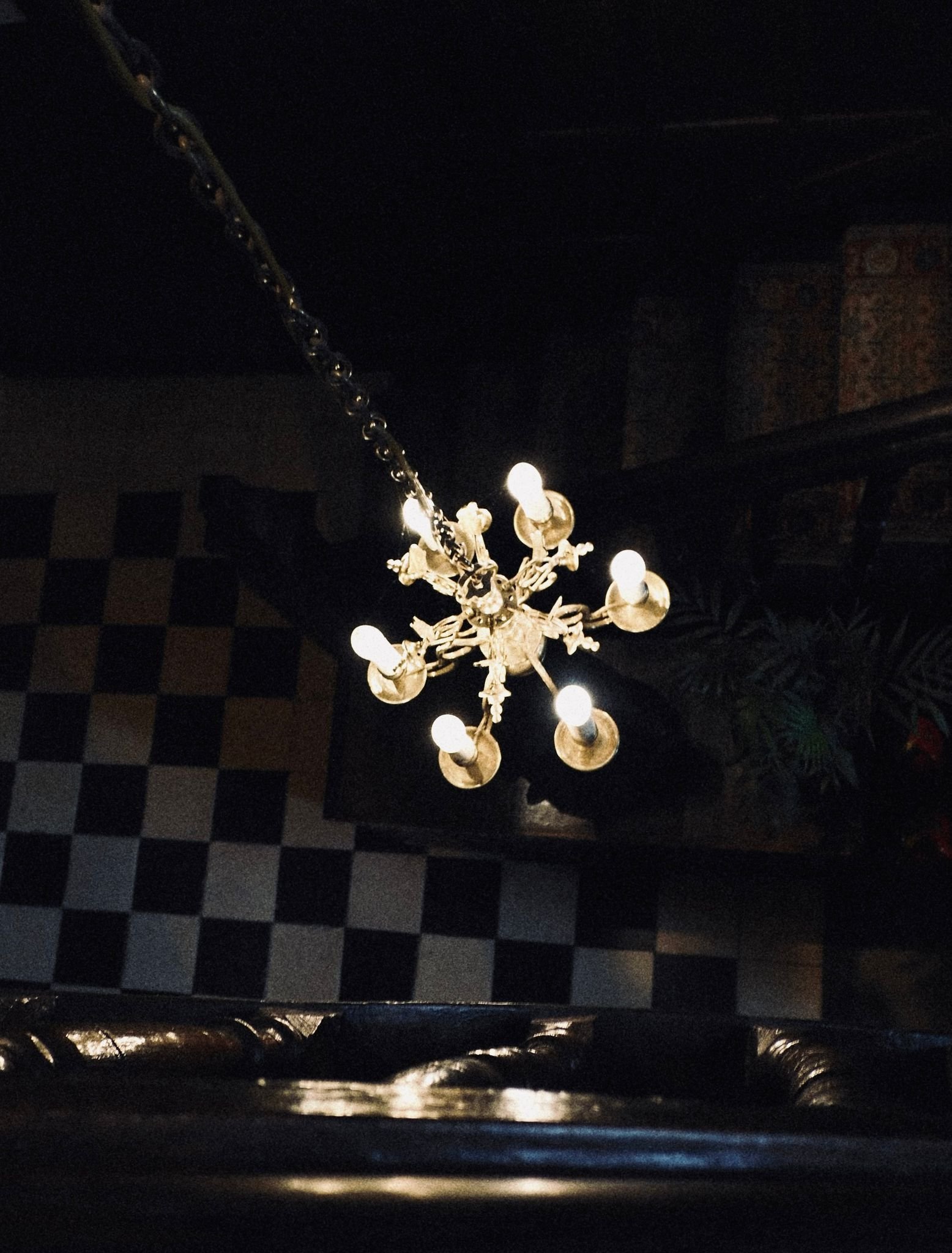 A chandelier hanging from the ceiling with multiple illuminated light bulbs, viewed from below, with a checkered black and white floor visible underneath.
