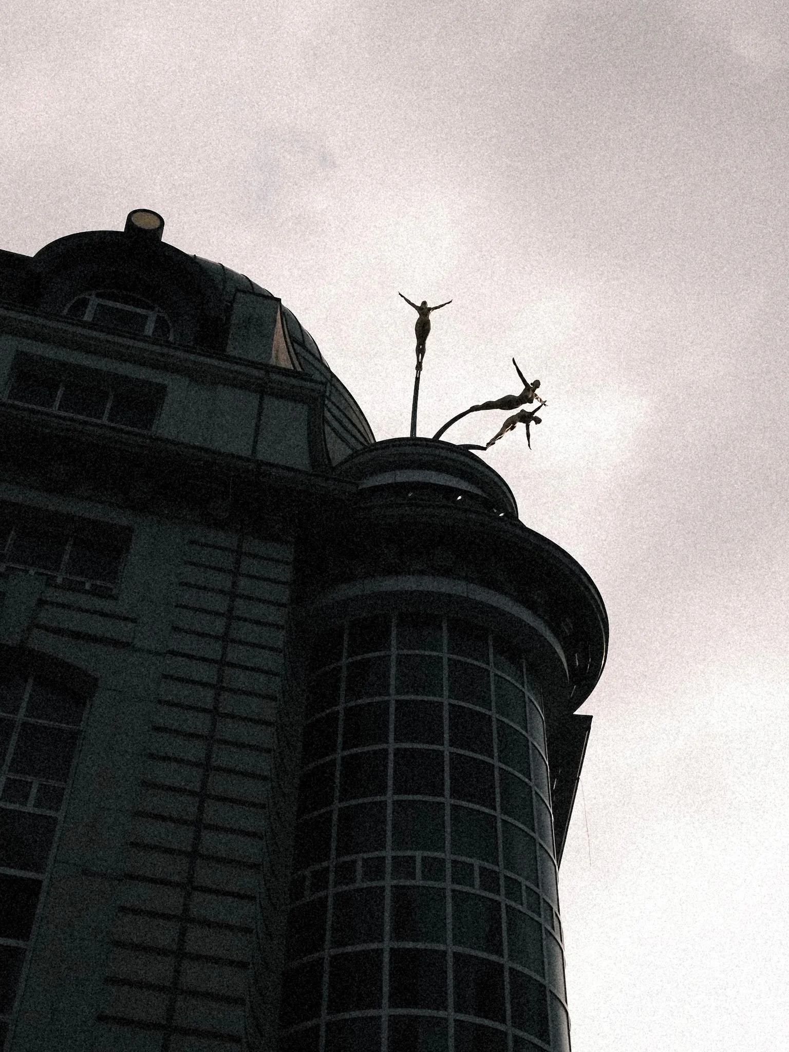 A tall building with a rounded glass tower at the corner, topped with a structure featuring three human sculptures with outstretched arms, resembling flying or floating.