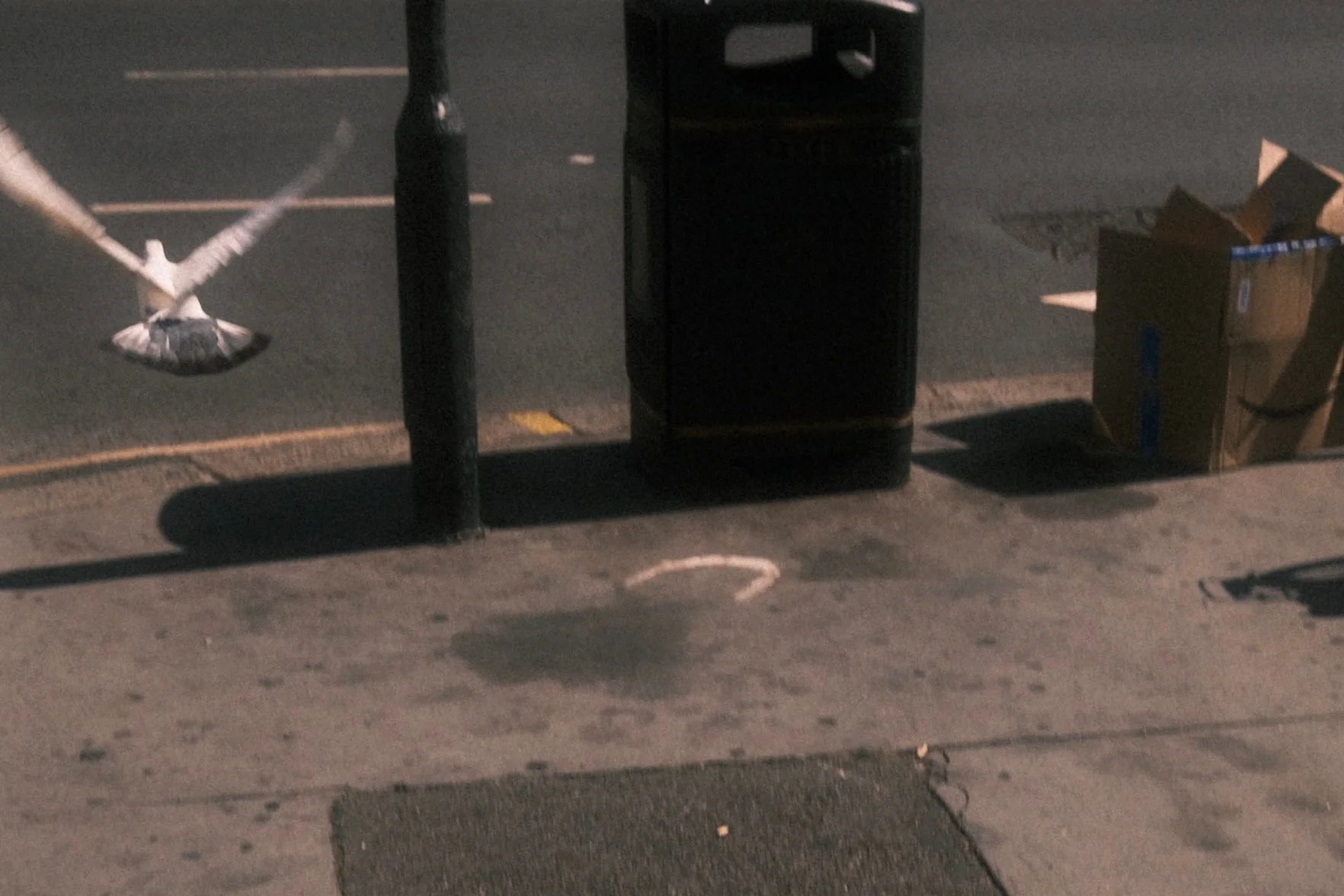 A sidewalk at night with a metal pole, trash bin, and cardboard boxes. There is a paper bag with a paper crane sculpture on the sidewalk.