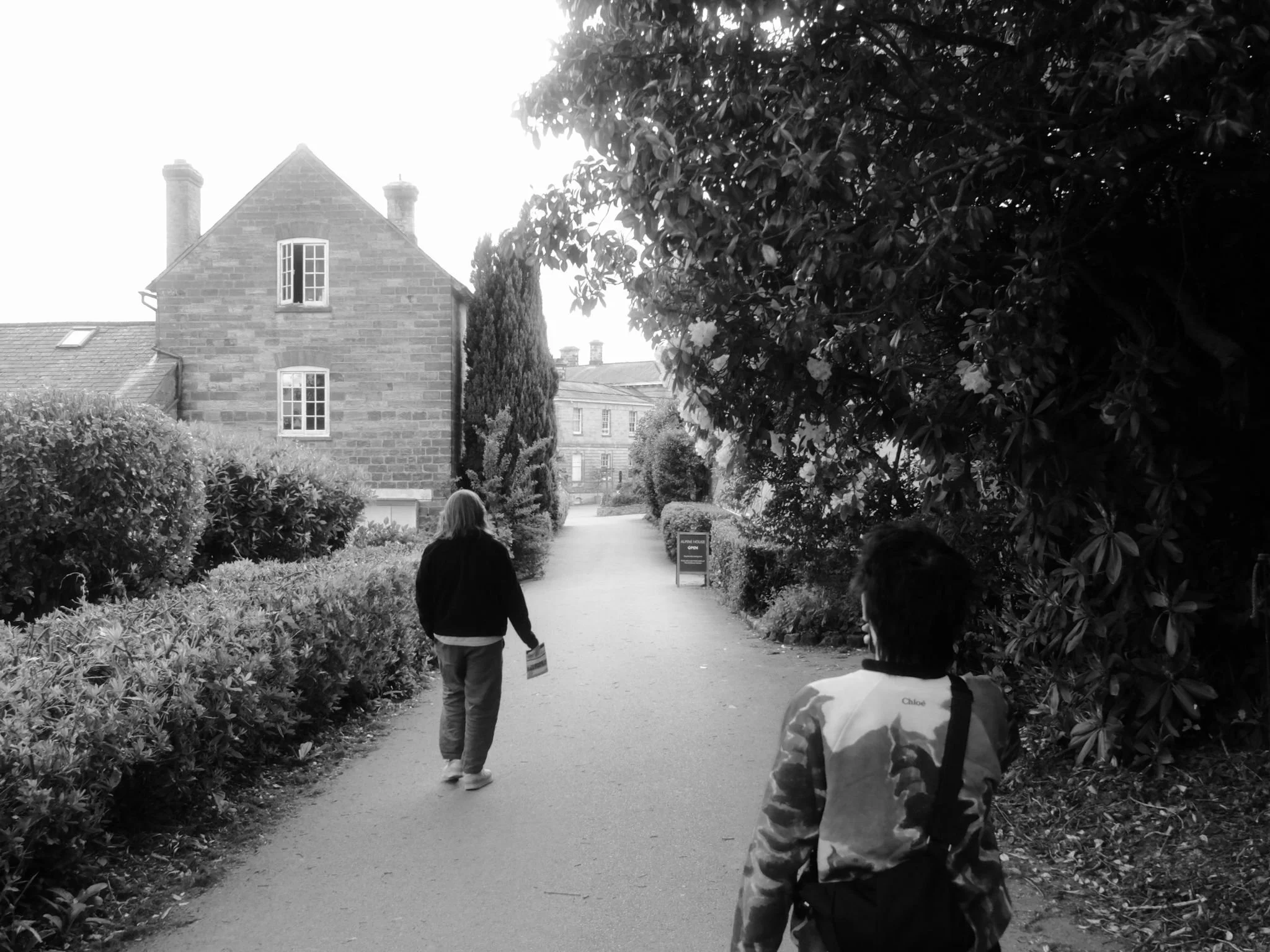 Two people walking along a paved pathway in a garden or park area, surrounded by bushes and trees, with a brick building in the background.