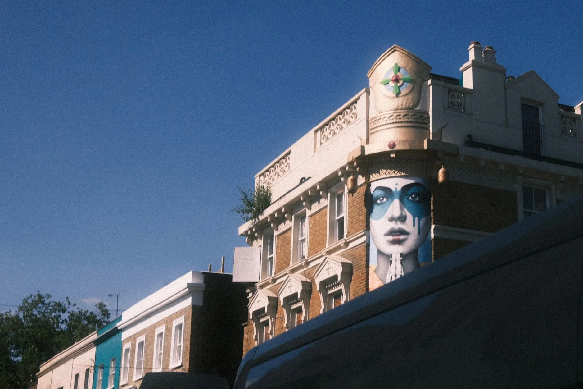 A building with a large mural of a woman with blue face paint and tears, and a decorative shield symbol on the top corner.