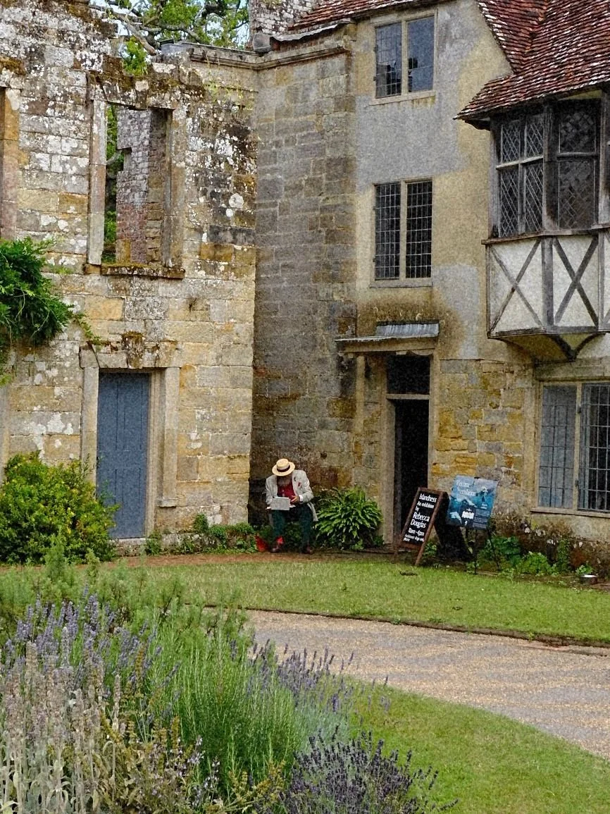 Person sitting on a bench reading a newspaper outside a historic stone building with a garden in the foreground.