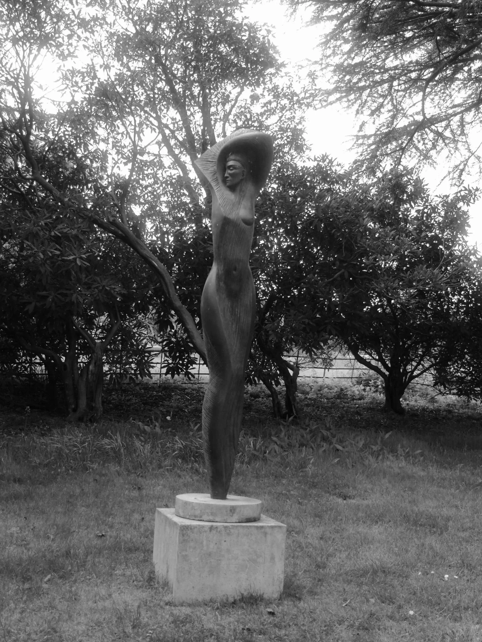 A sculpture of a woman with a tall, elongated body and flowing hair, standing on a circular base in a garden surrounded by trees.