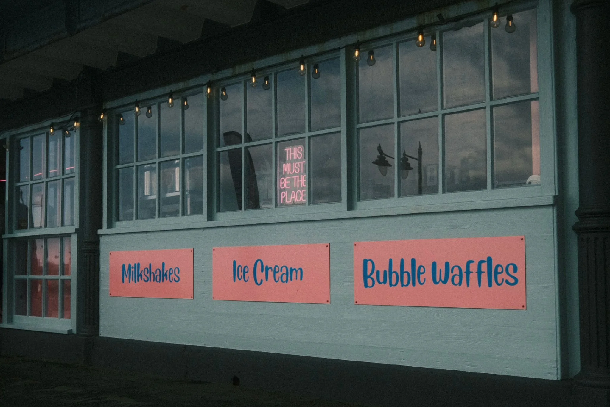 A storefront with three pink signs advertising Milkshakes, Ice Cream, and Bubble Waffles. Above, a window with a pink neon sign that reads 'This must be the place' is visible, along with string lights hanging from the roof.