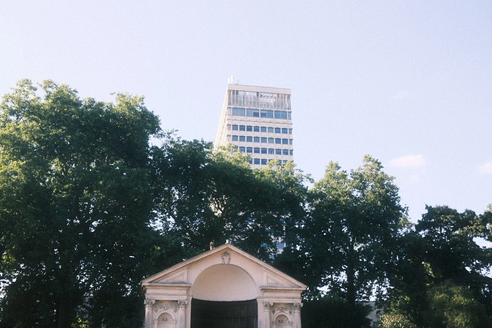 Tall modern skyscraper with a slanted roof seen above a hillside with trees and a classical white temple or small building in the foreground.