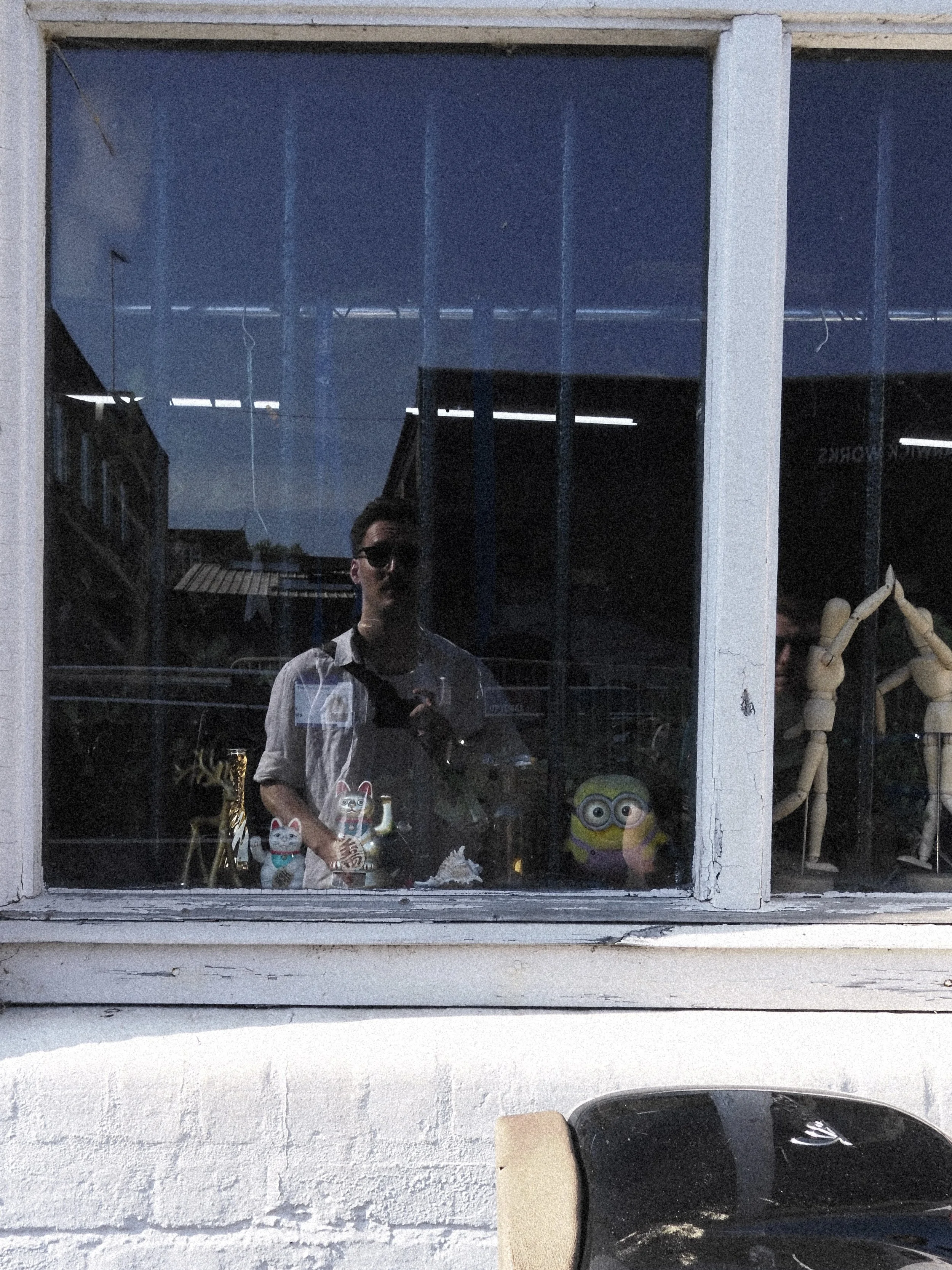 Reflection of a man taking a photo through a window with various figurines and decorations visible inside the building.
