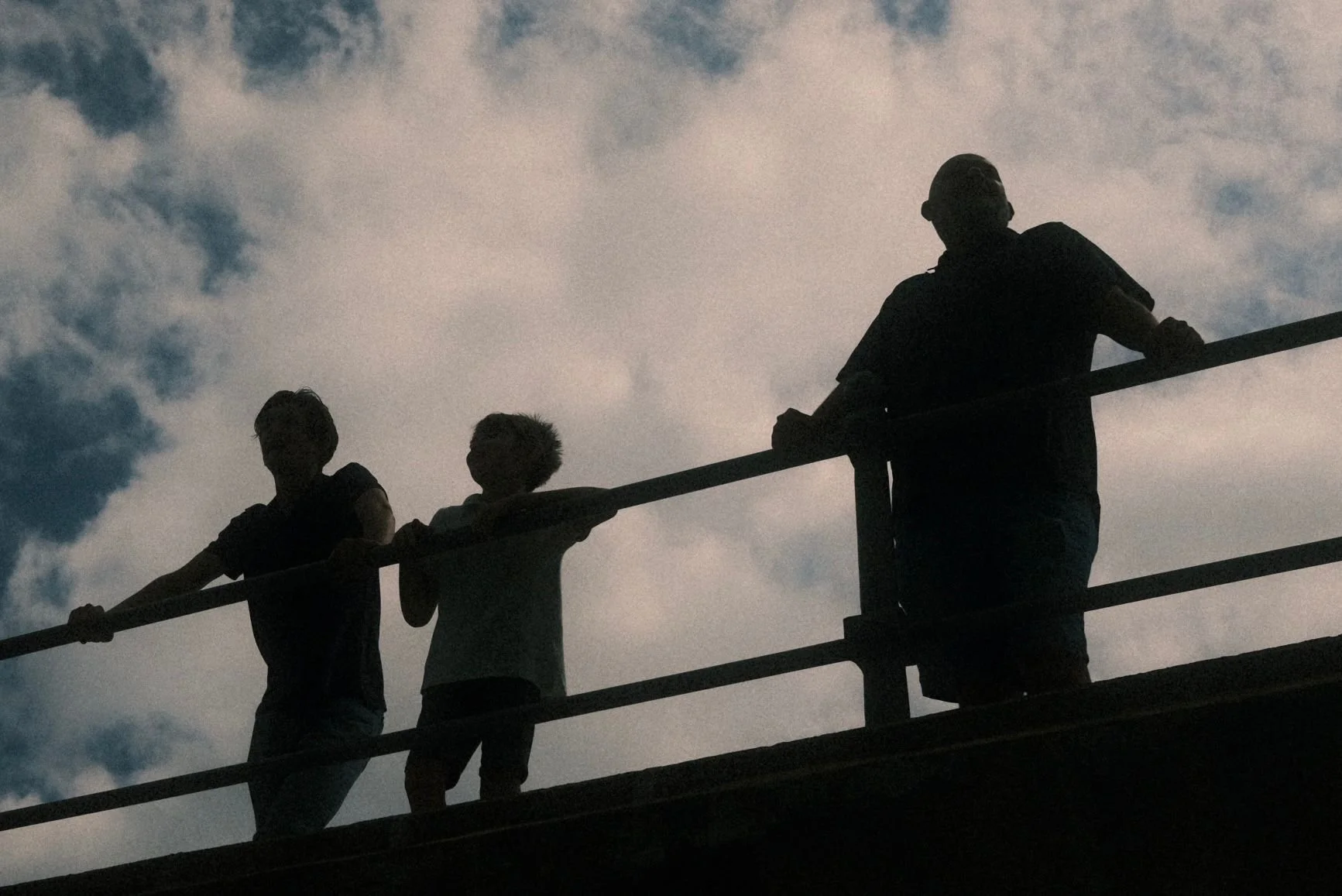 Silhouettes of three people, two children and an adult, standing on a balcony or platform with railing, against a cloudy sky during daytime.