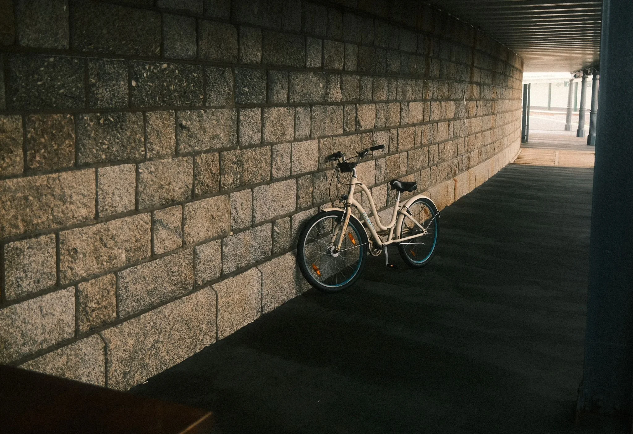 A white bicycle is parked against a brick wall in a covered walkway, with a shaded area and columns visible in the background.