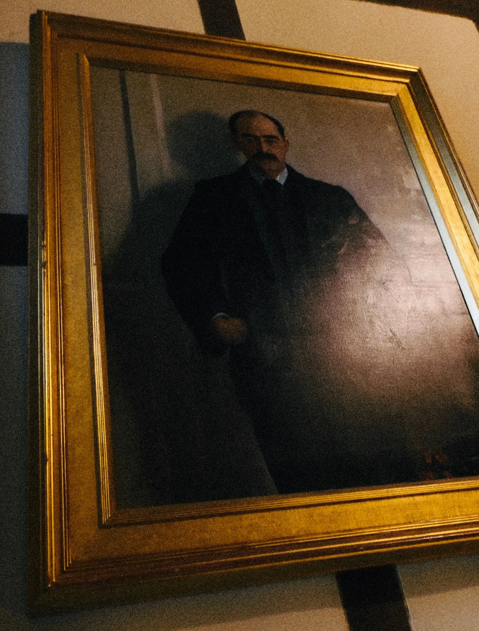 A portrait painting of a man with a mustache, wearing a dark suit and tie, inside a gold frame, hung on a wall.