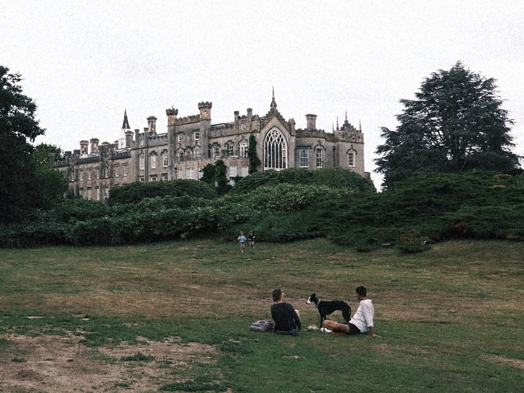 People sitting on grass in front of a large castle-like building with gothic windows and turrets, surrounded by trees and shrubs.