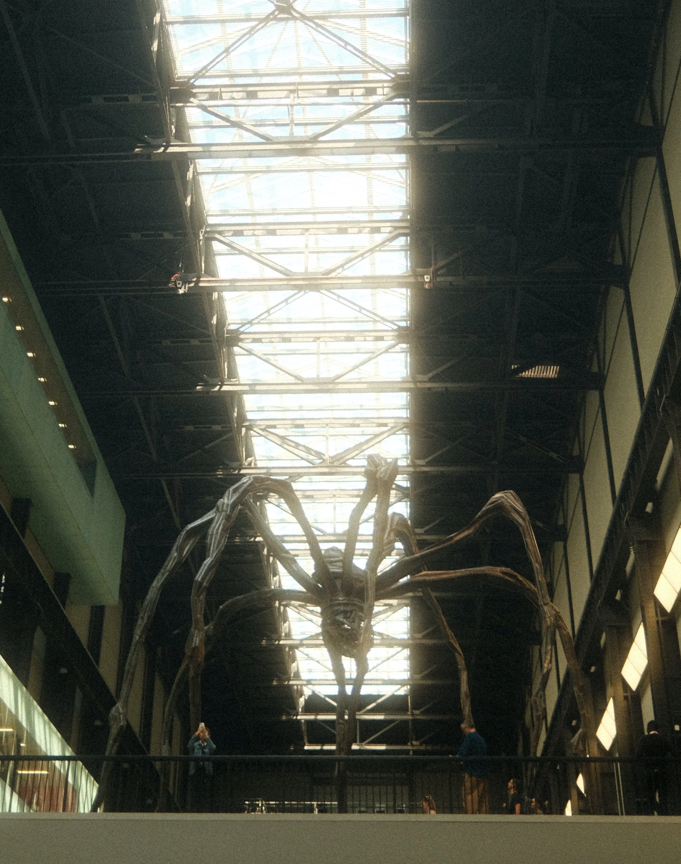An indoor museum displaying a large spider sculpture and a dinosaur skeleton. The spider appears to be made of metallic materials and is prominently placed in the center.