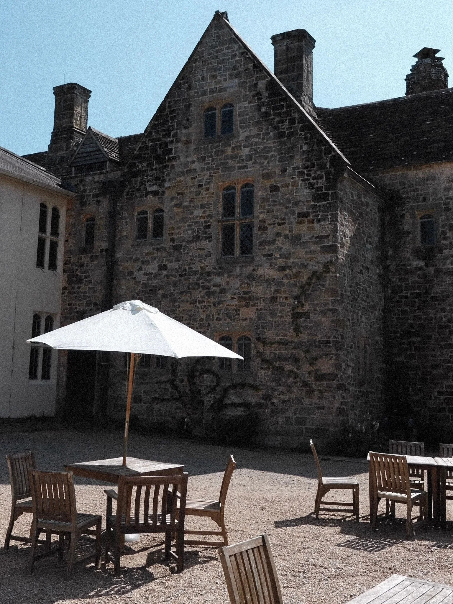 Outdoor seating area with wooden tables and chairs, one with a white umbrella, in front of a large stone historic building with multiple stories, chimneys, and arched windows, under a clear blue sky.