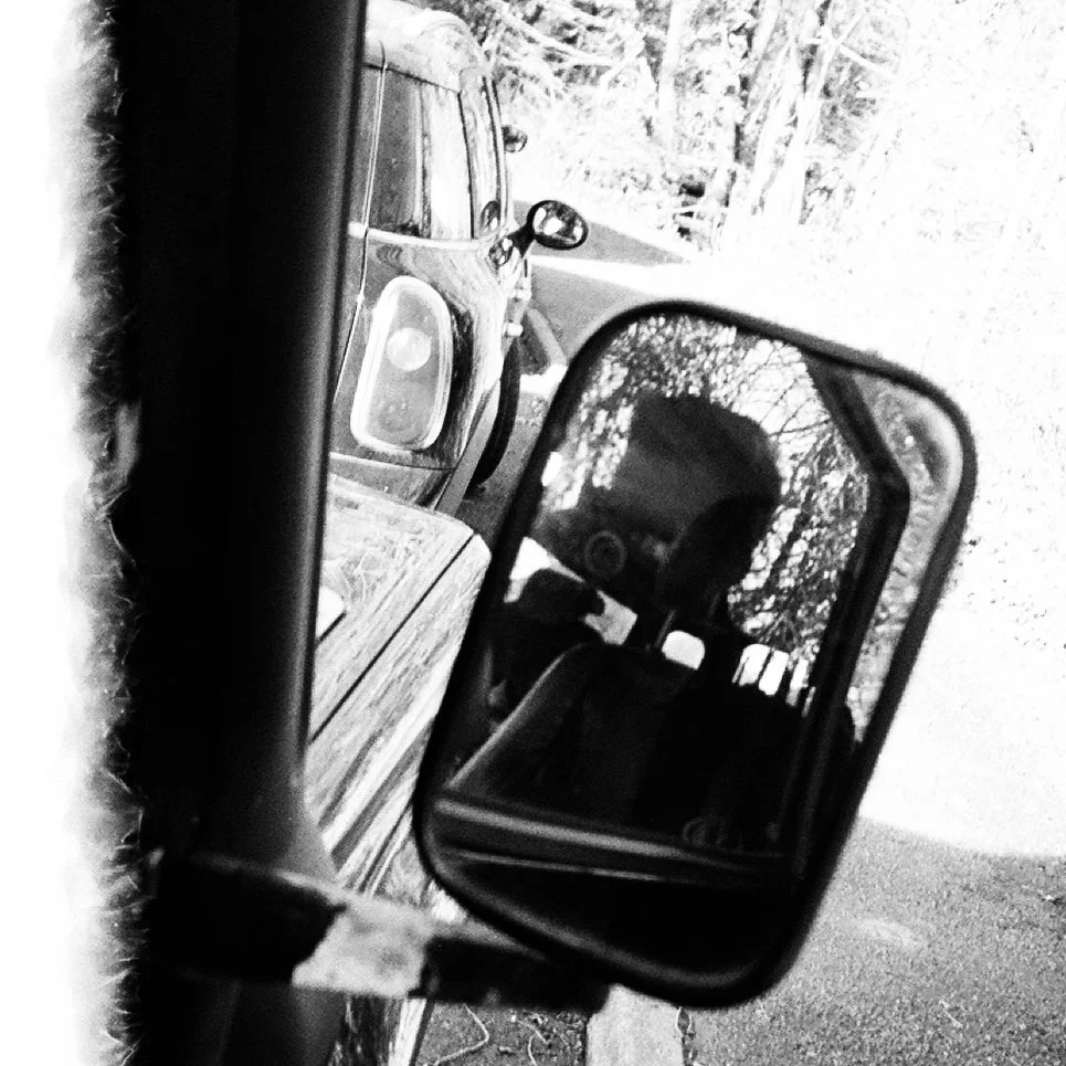 Black and white photo of a car's side mirror reflecting the interior of the vehicle and a wooded area behind the car, with another vehicle parked alongside.