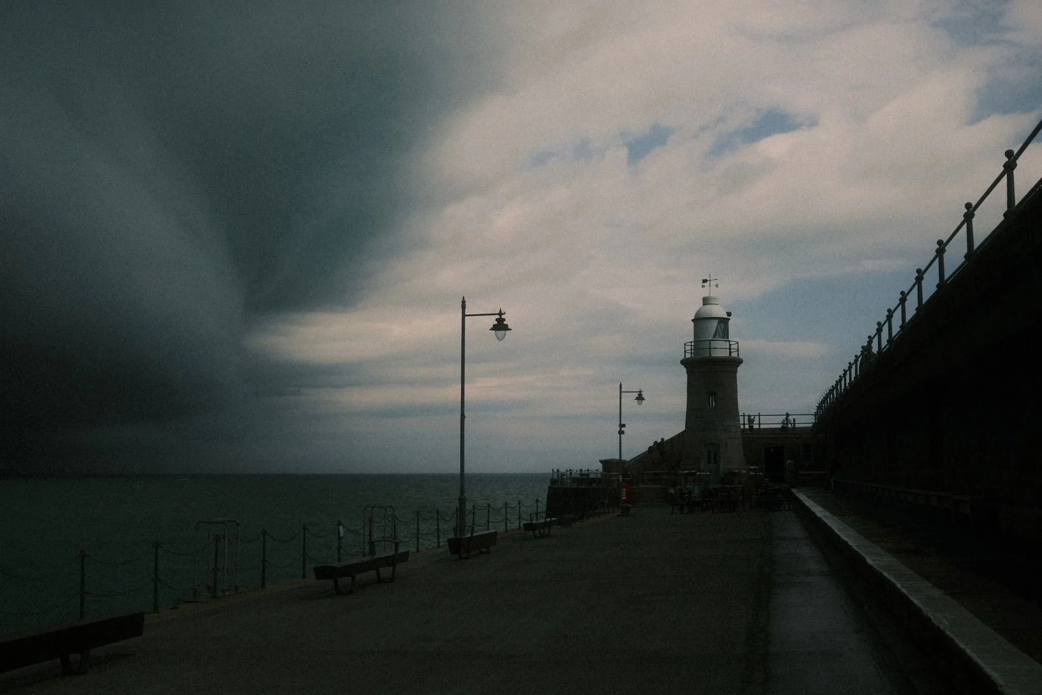 A lighthouse on a pier with benches, lampposts, and a railing, under a stormy sky with dark clouds on the left and some light clouds on the right.