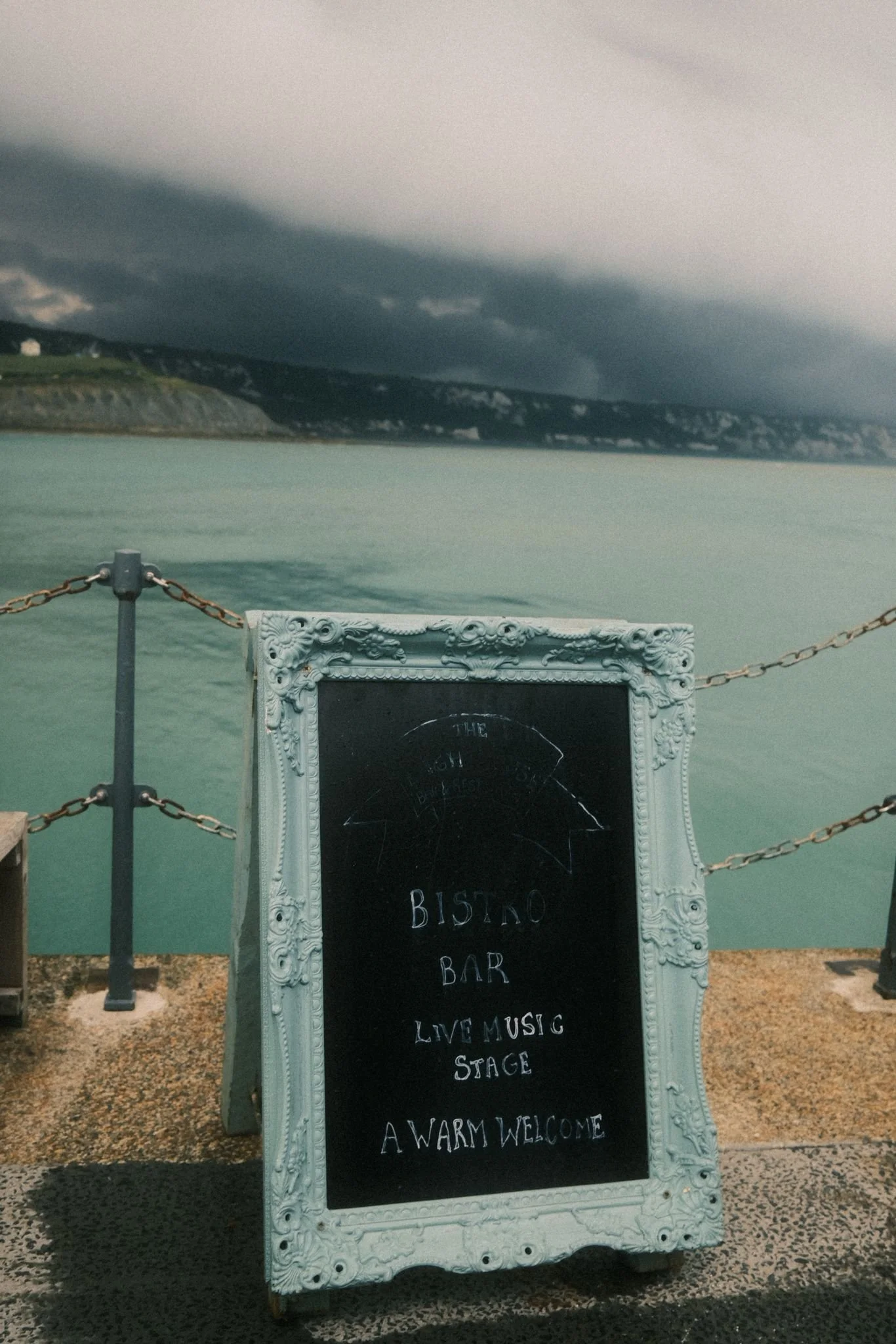A chalkboard sign on a pier with water and hills in the background, advertising a bistro bar with live music and a welcoming atmosphere.