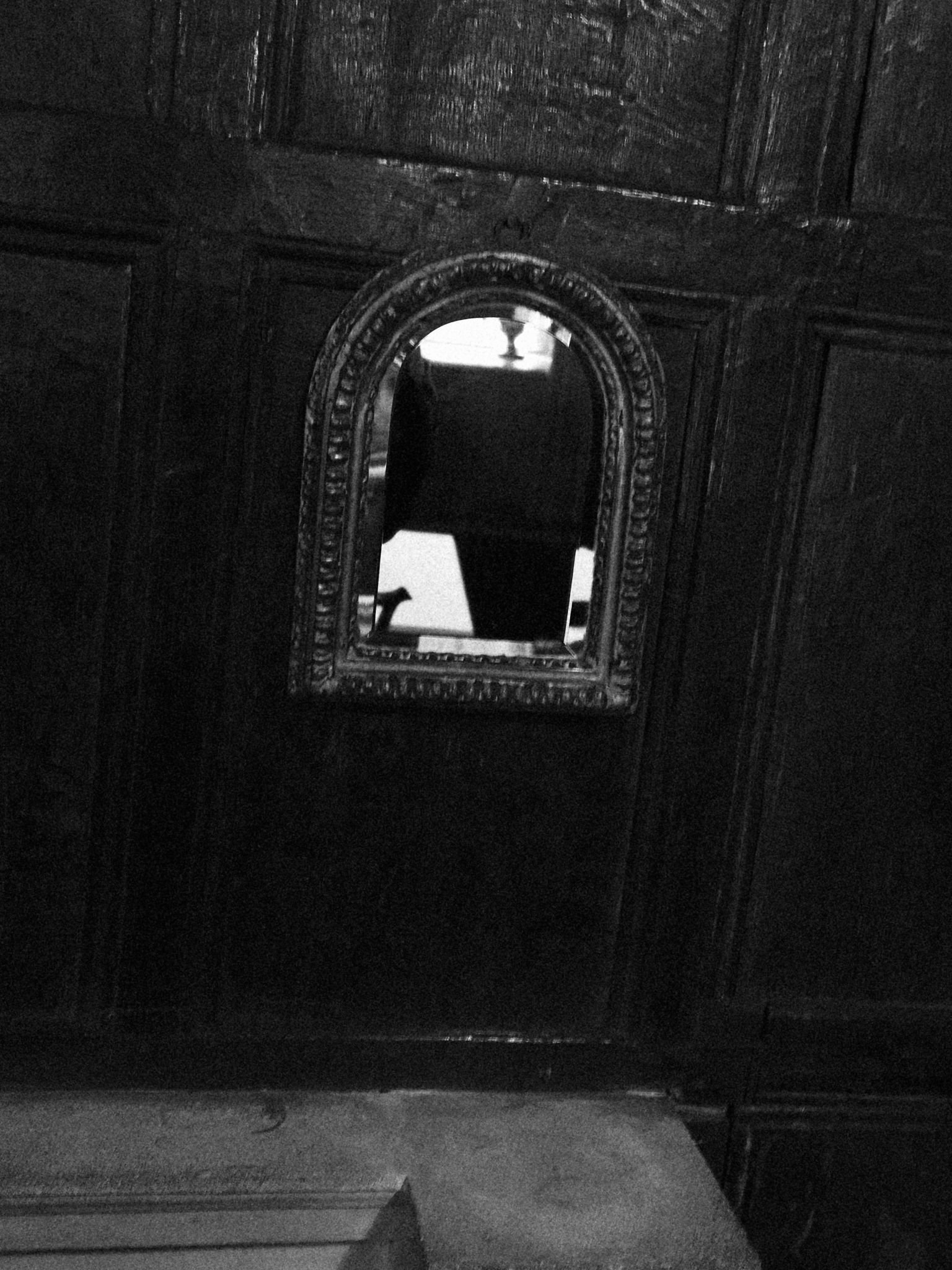 A black and white photo of a small arched mirror embedded in a dark wooden wall or cabinet, reflecting part of an object or furniture and a window or light source.