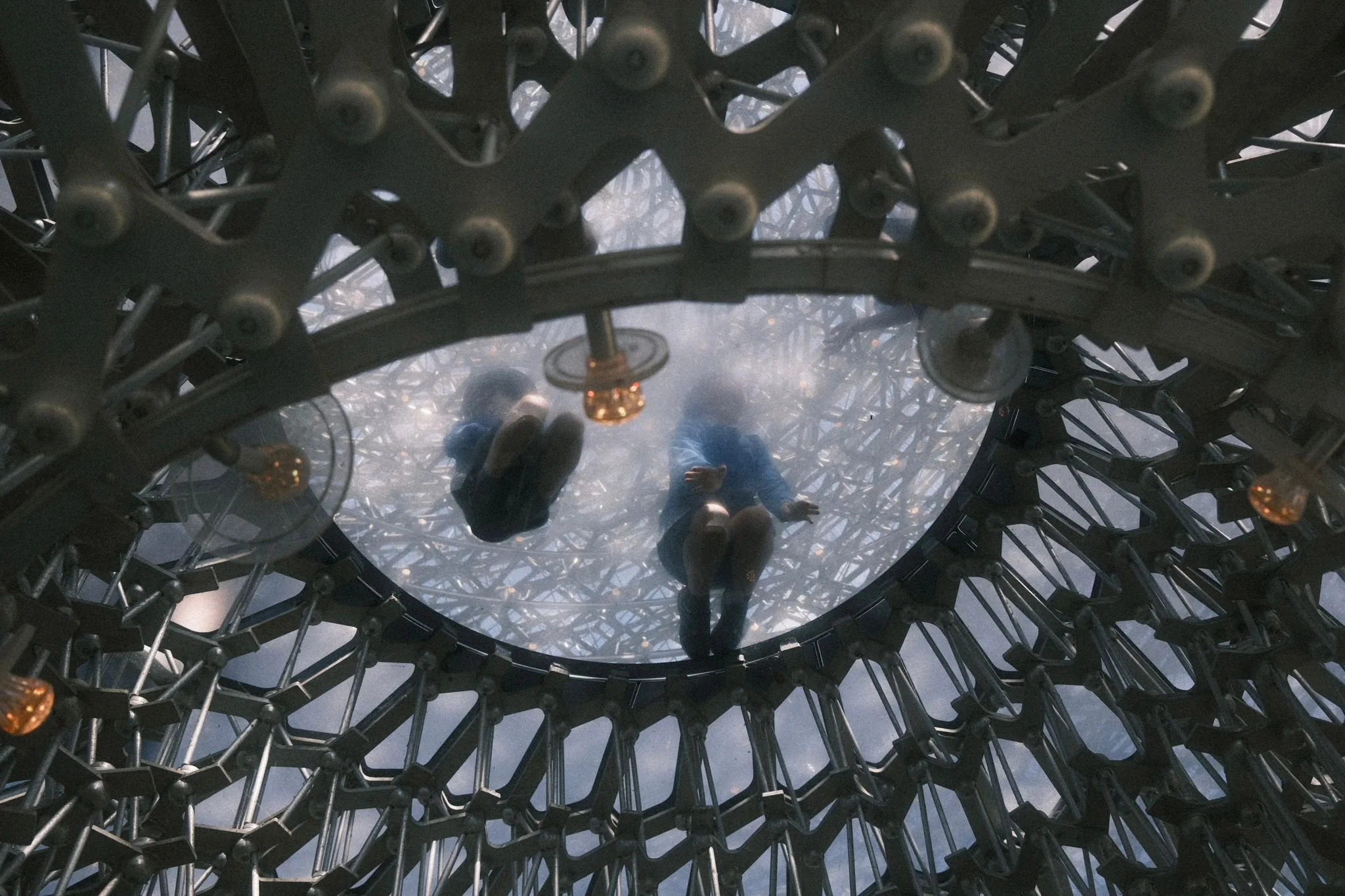 Two people are reflected in a large, curved mirror with a metallic, web-like frame. The mirror is elevated, and the people are looking down at it.