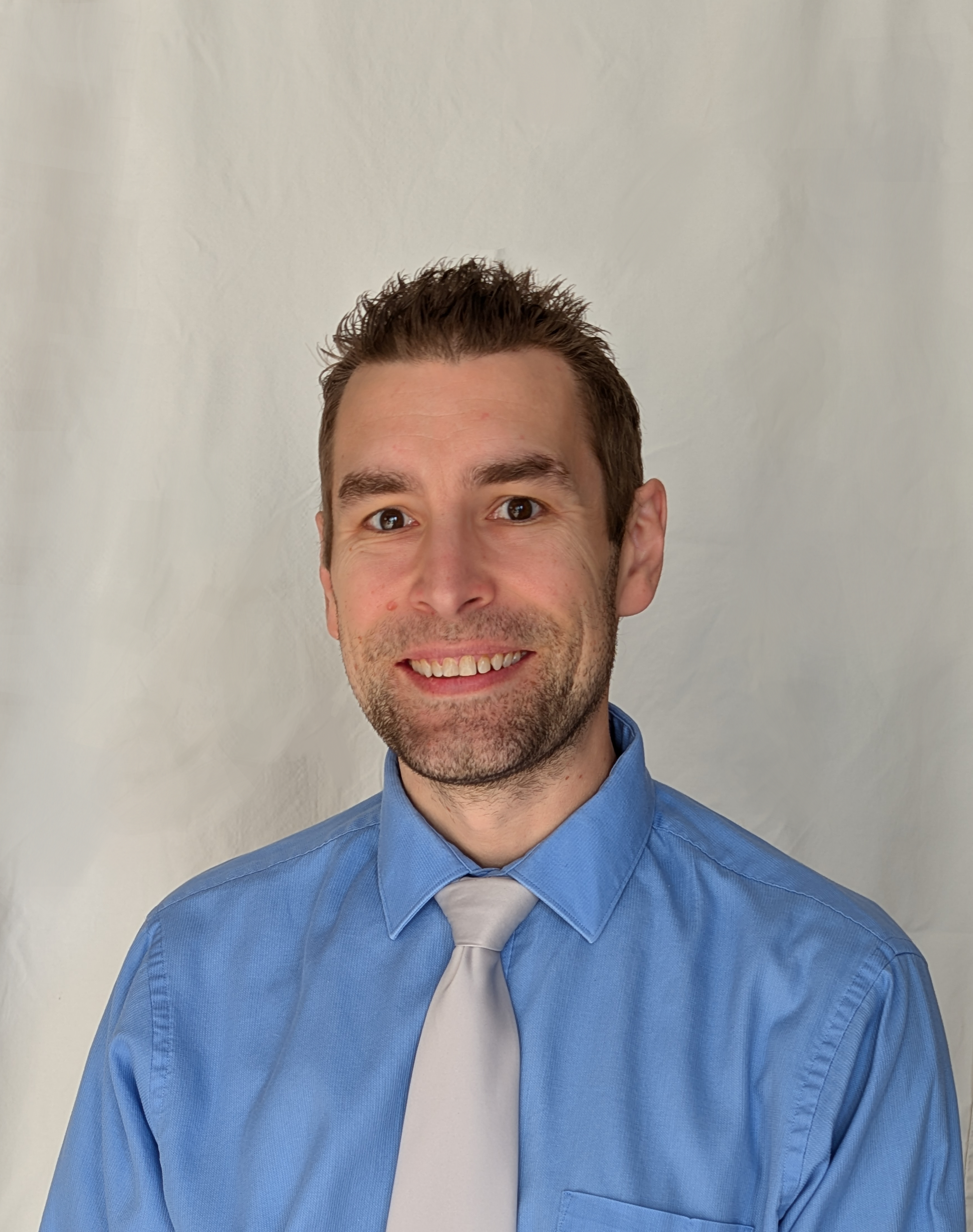 Portrait of a smiling man with short brown hair, wearing a blue dress shirt and a light gray tie, set against a plain light-colored background.