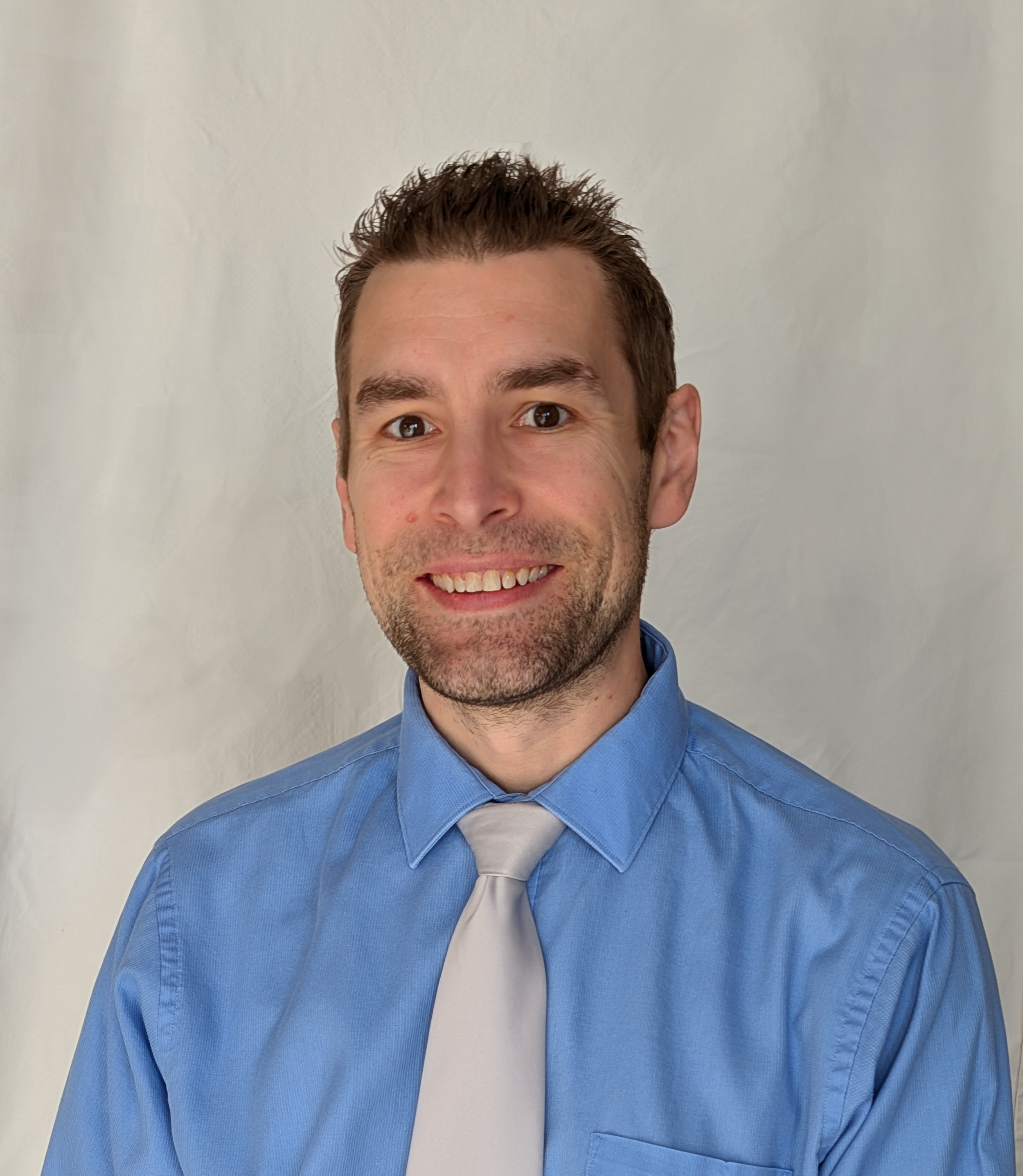 Headshot of a smiling man with light skin, short brown hair, wearing a light blue dress shirt and a light-colored tie, against a plain neutral background.
