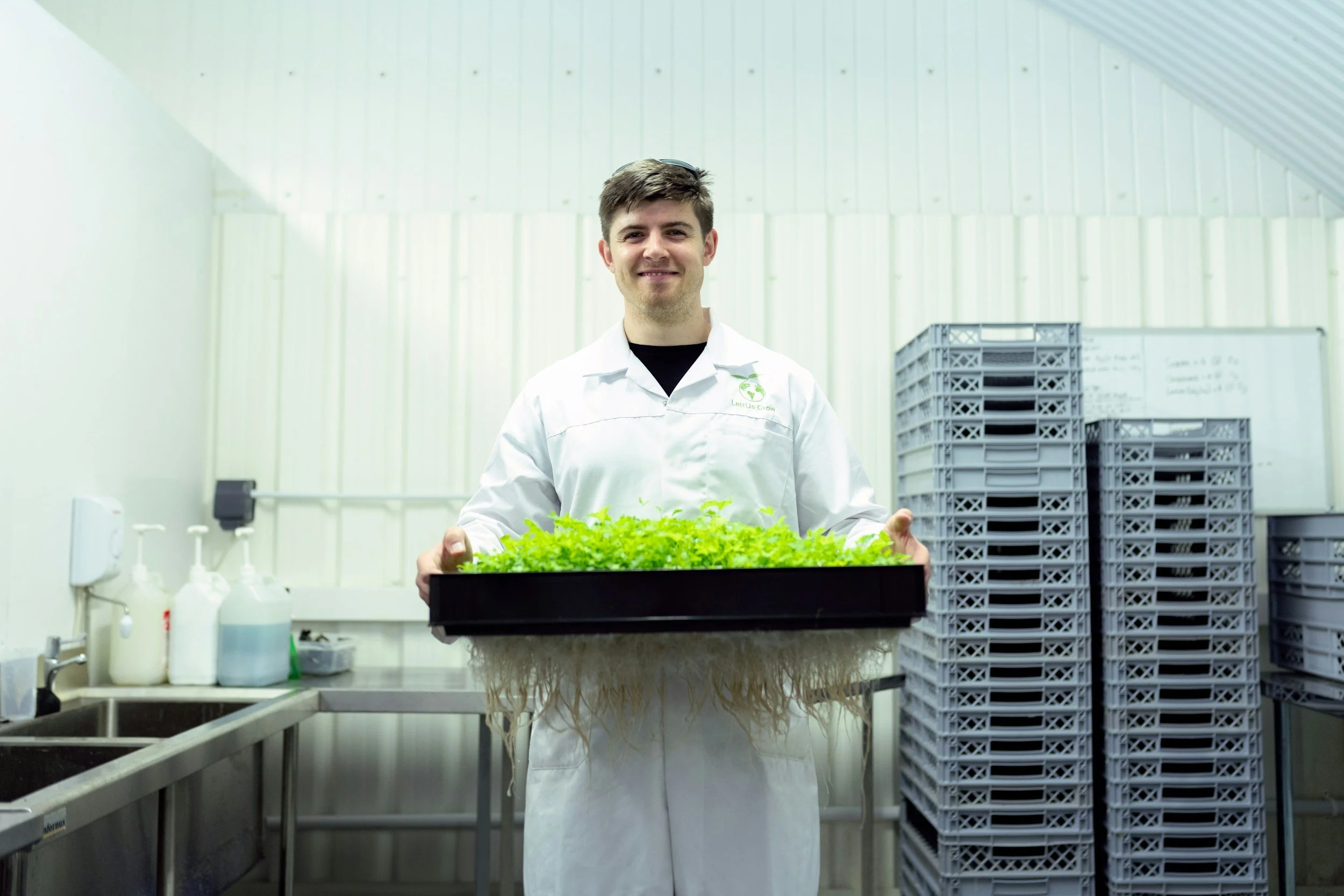 A man in a white lab coat holding a tray of green seedlings in a laboratory or cultivation facility utilizing Biocat PST Phage soil technology