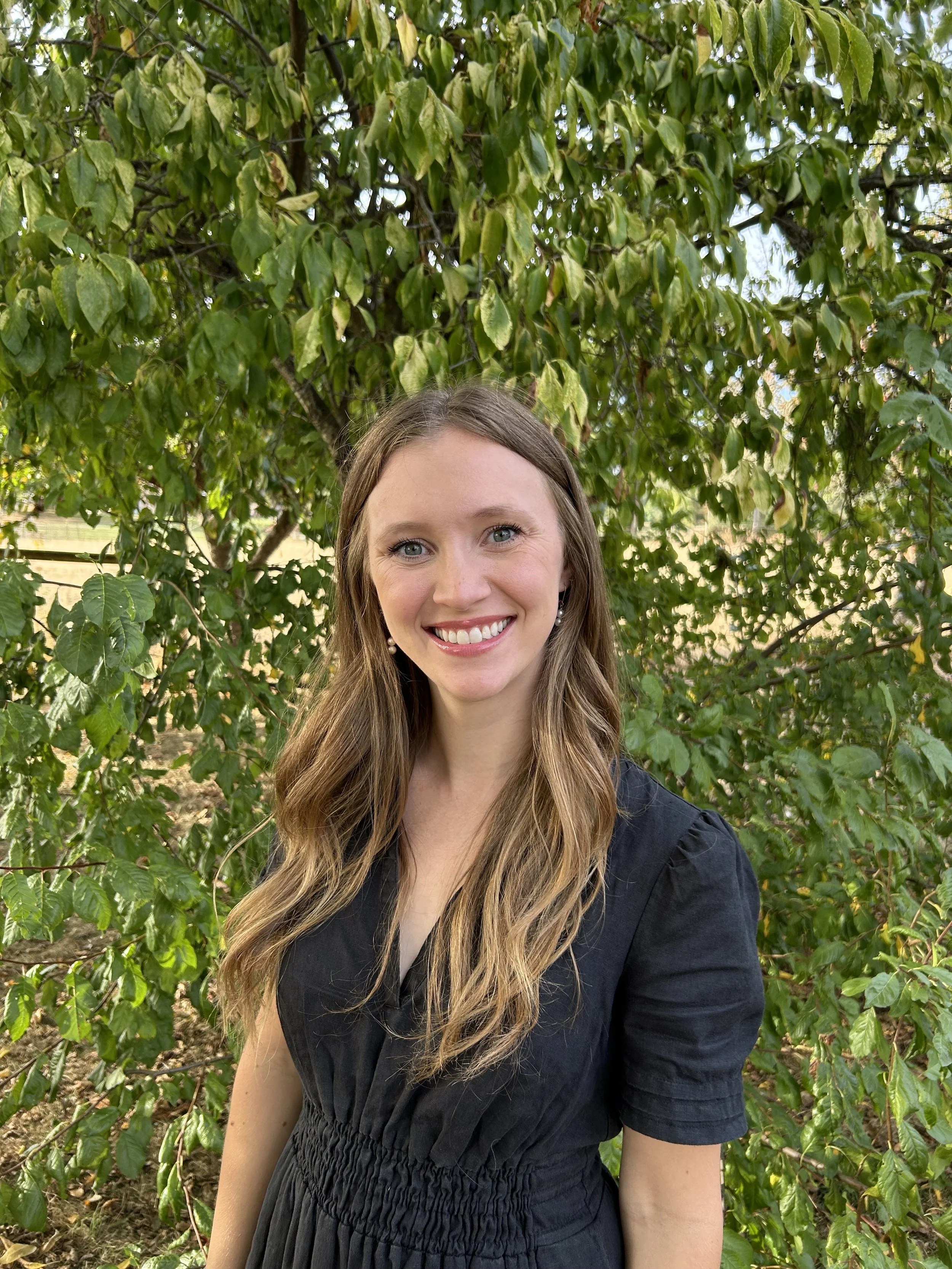 A woman with long, wavy blonde hair and blue eyes smiling outdoors in front of green leafy trees, wearing a black dress.
