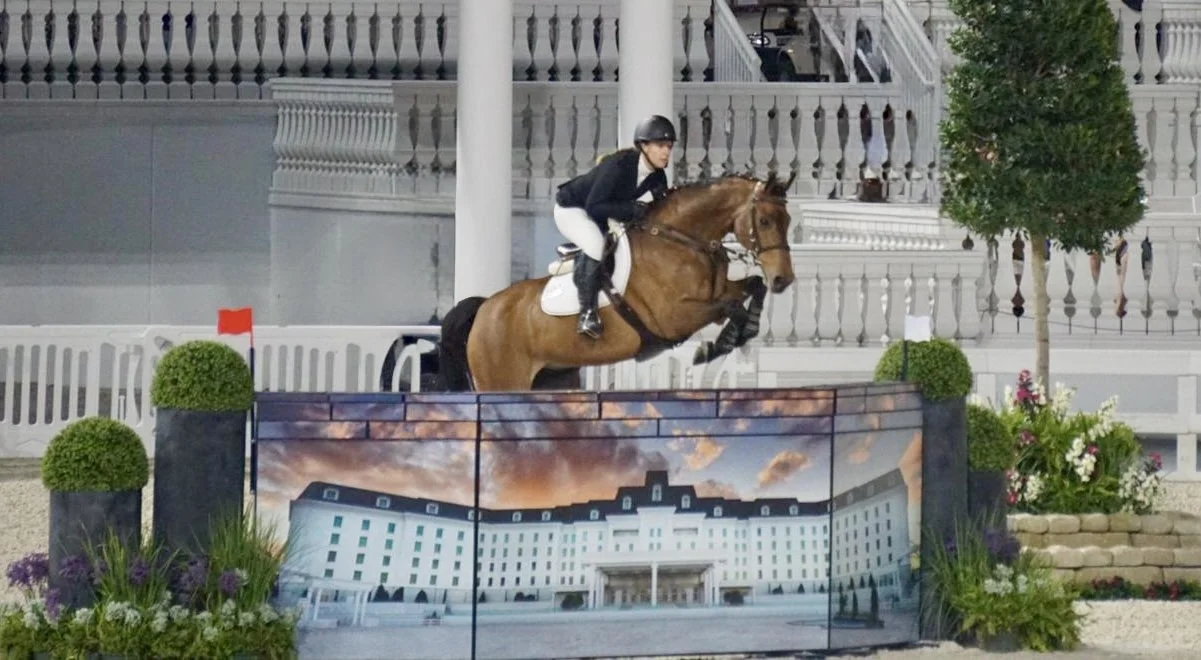 Bryn Sadler riding Cinven CR jumping the wall with the grand hotel printed on it in the Grand Prix at WEC Ocala, Florida