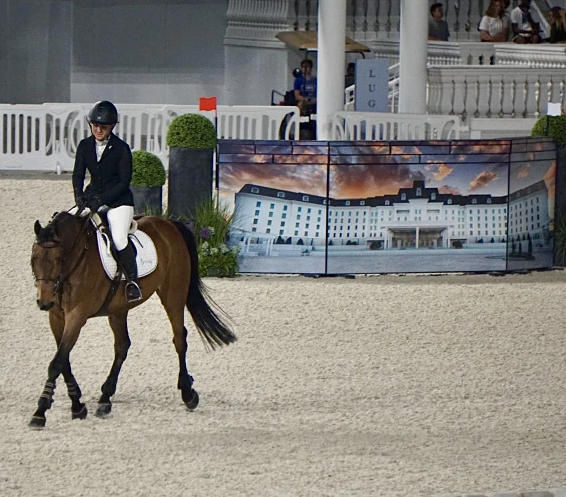 Bryn Sadler riding Cinven CR trotting in the ring at WEC Ocala with the Grand Hotel wall jump backdrop