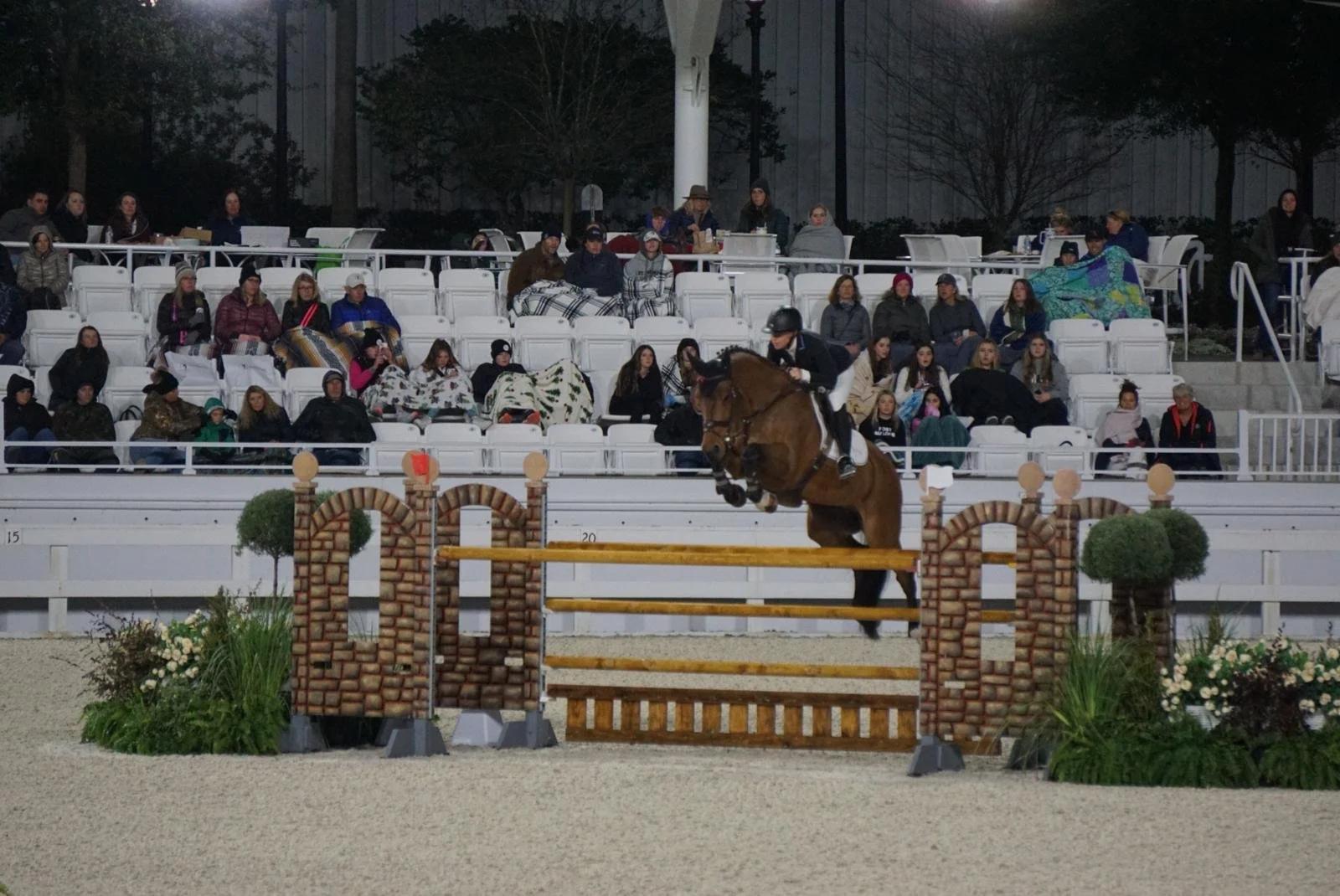 A rider on a galloping horse clearing a jump in an equestrian competition, with an audience watching in the background.