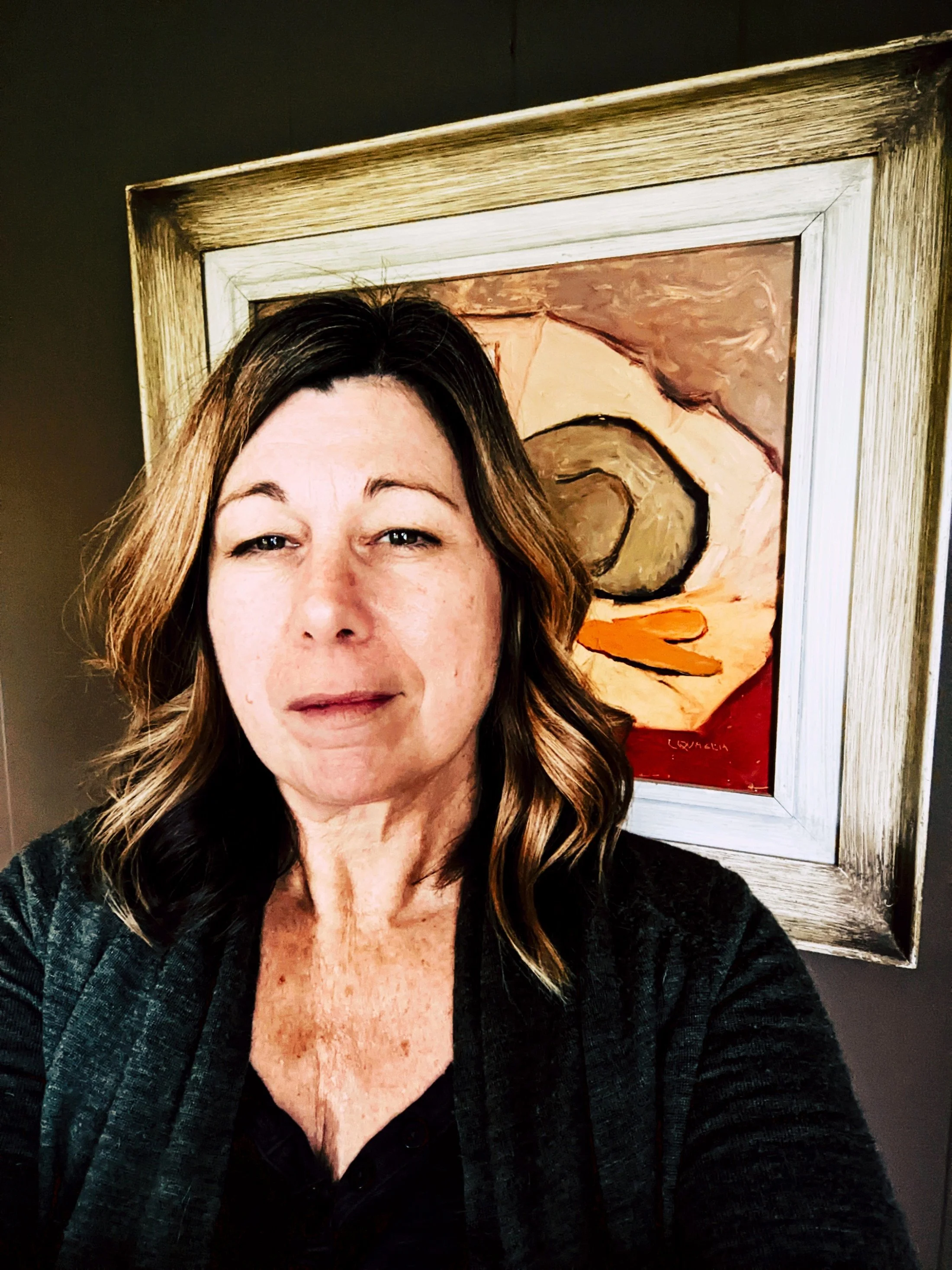 A person with wavy brown hair and a black top taking a selfie in front of an abstract portrait painting with earth tones and bold strokes.