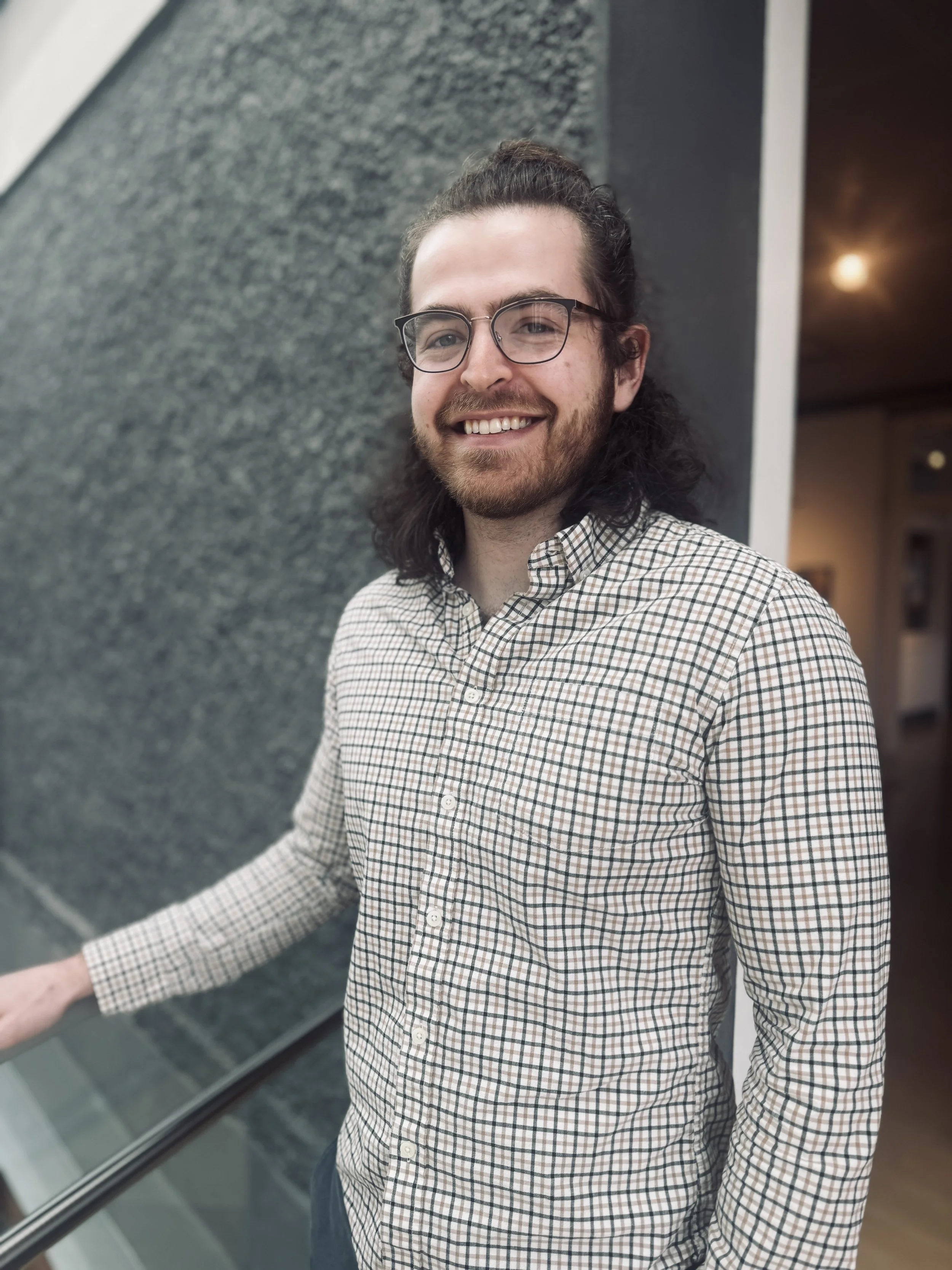 A person with long dark hair, beard, and glasses wearing a checkered shirt, smiling, standing near a textured wall in an indoor setting.