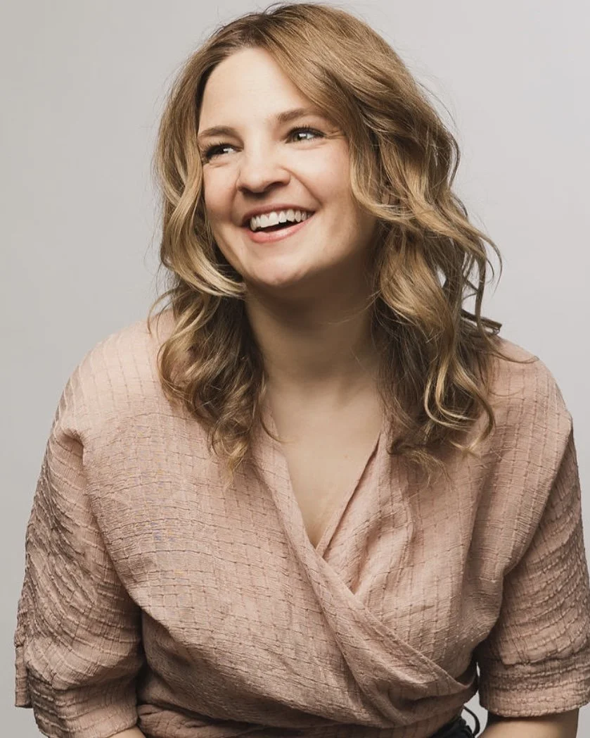 A person with wavy blonde hair, smiling and looking to her right, against a plain background, wearing a light pink textured blouse.